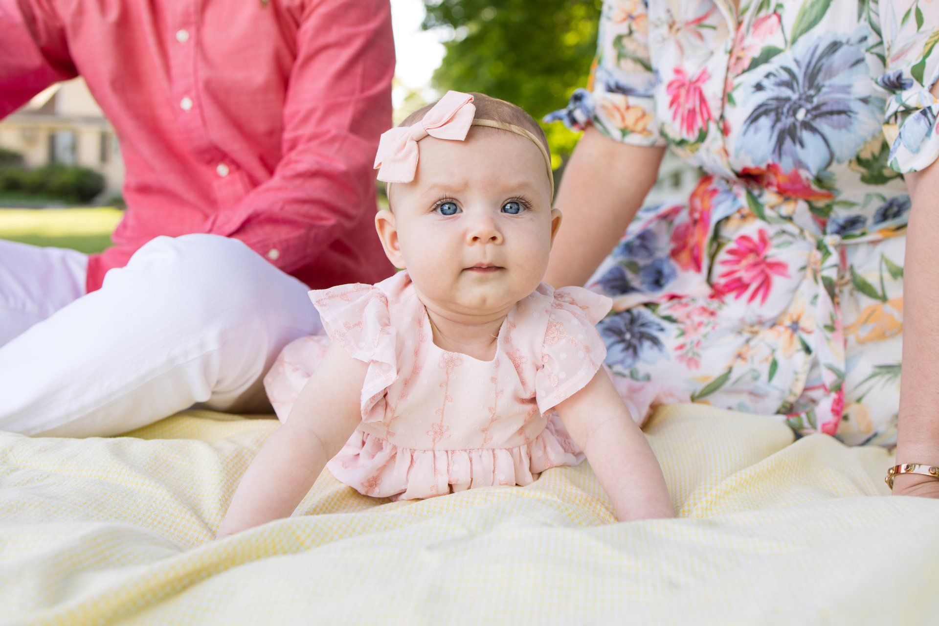 Baby in pink dress sitting on a blanket outdoors beside two adults in floral clothing