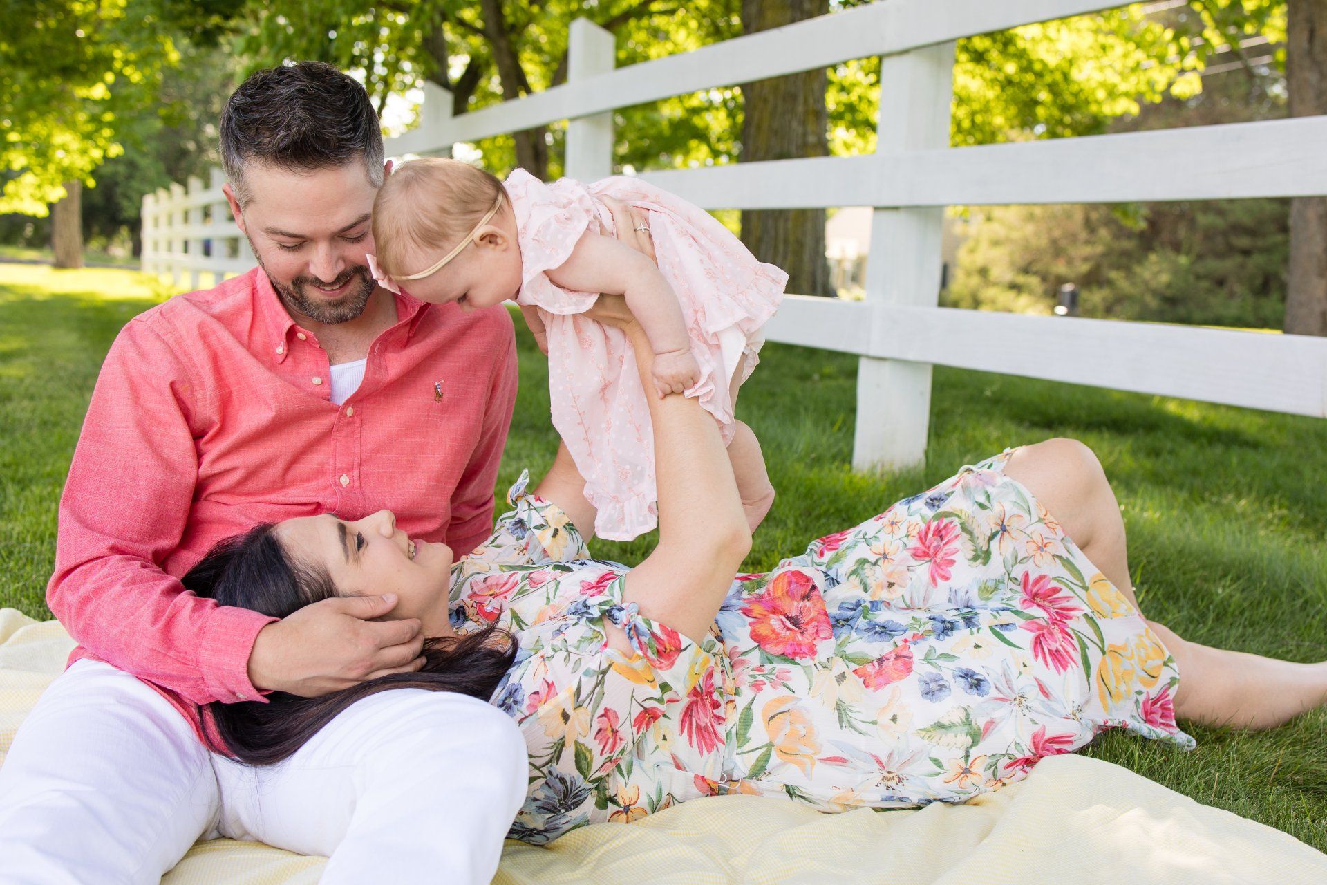 Family lying on a blanket outdoors, smiling at a baby being lifted in the air