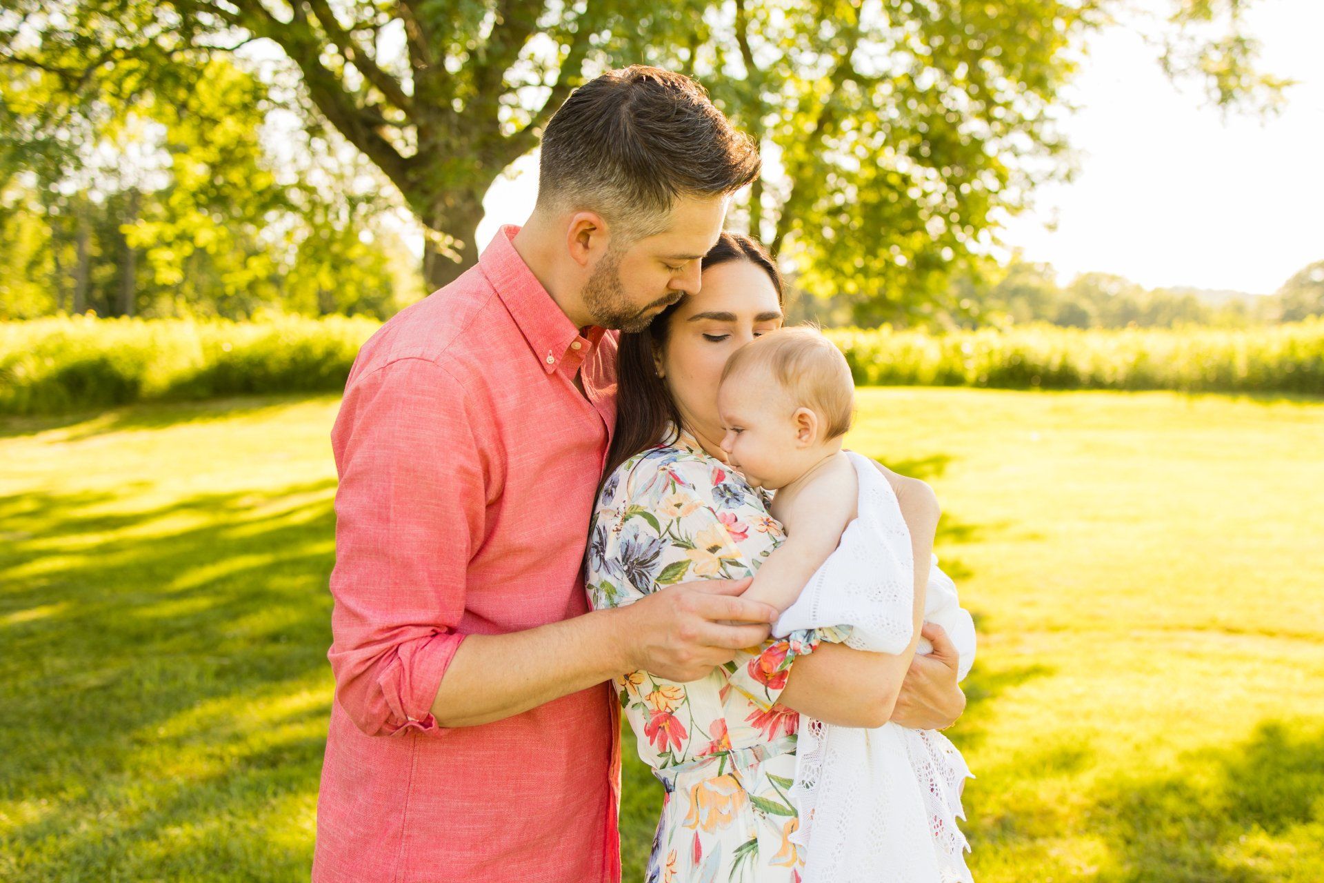 Adult holding and kissing a baby in a sunny park with green grass and trees