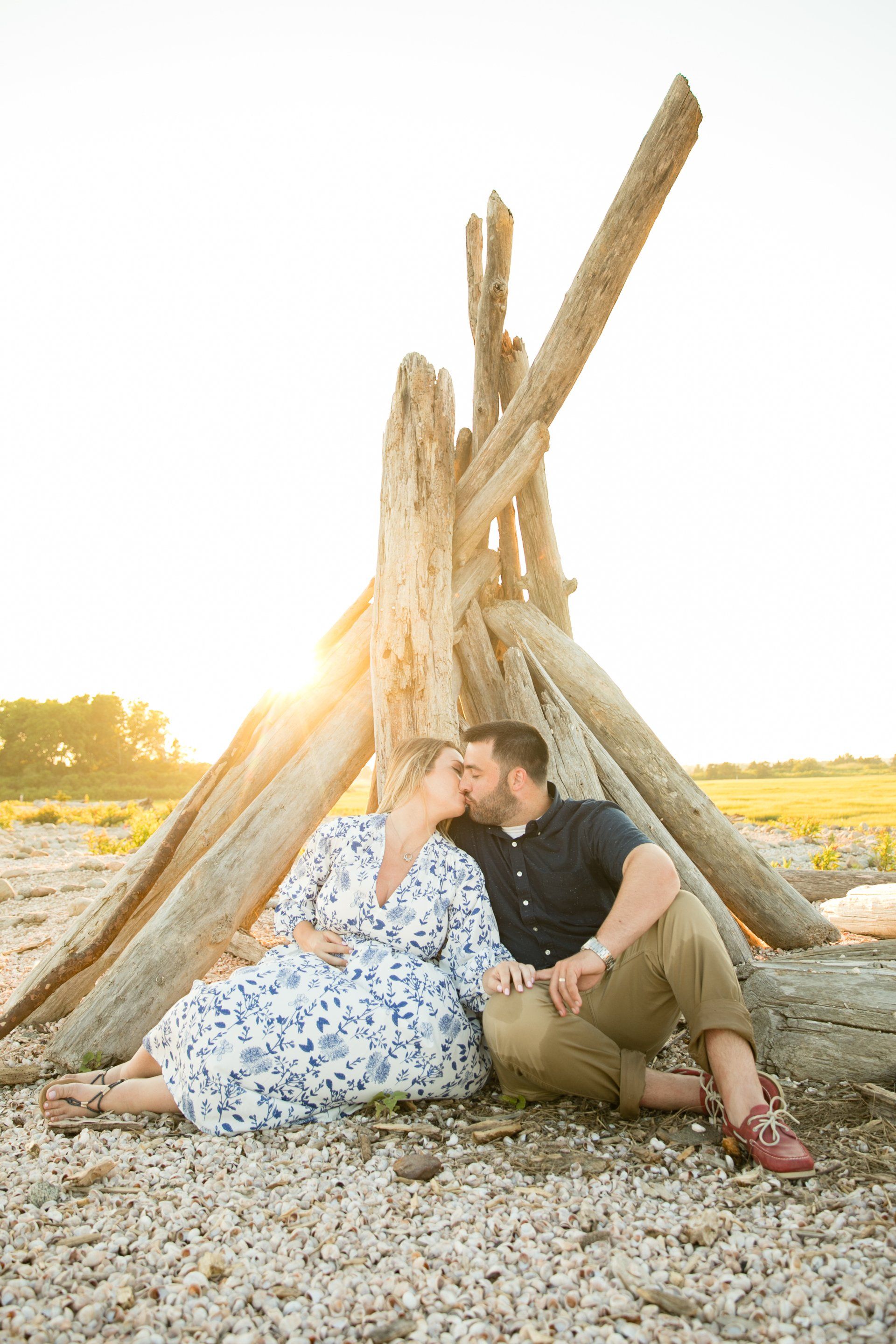 Couple sitting and leaning against a driftwood teepee on a rocky beach at sunset