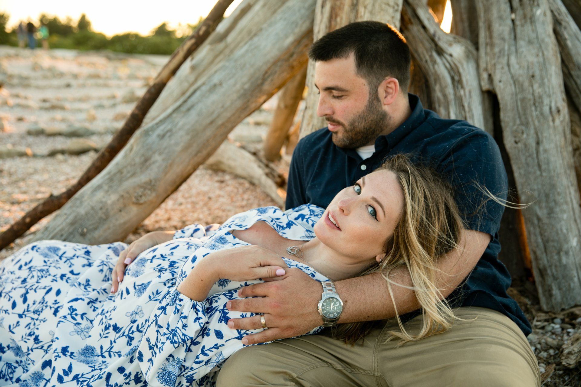 Couple relaxing outdoors by a rustic wooden structure at sunset, woman reclined in man’s arms