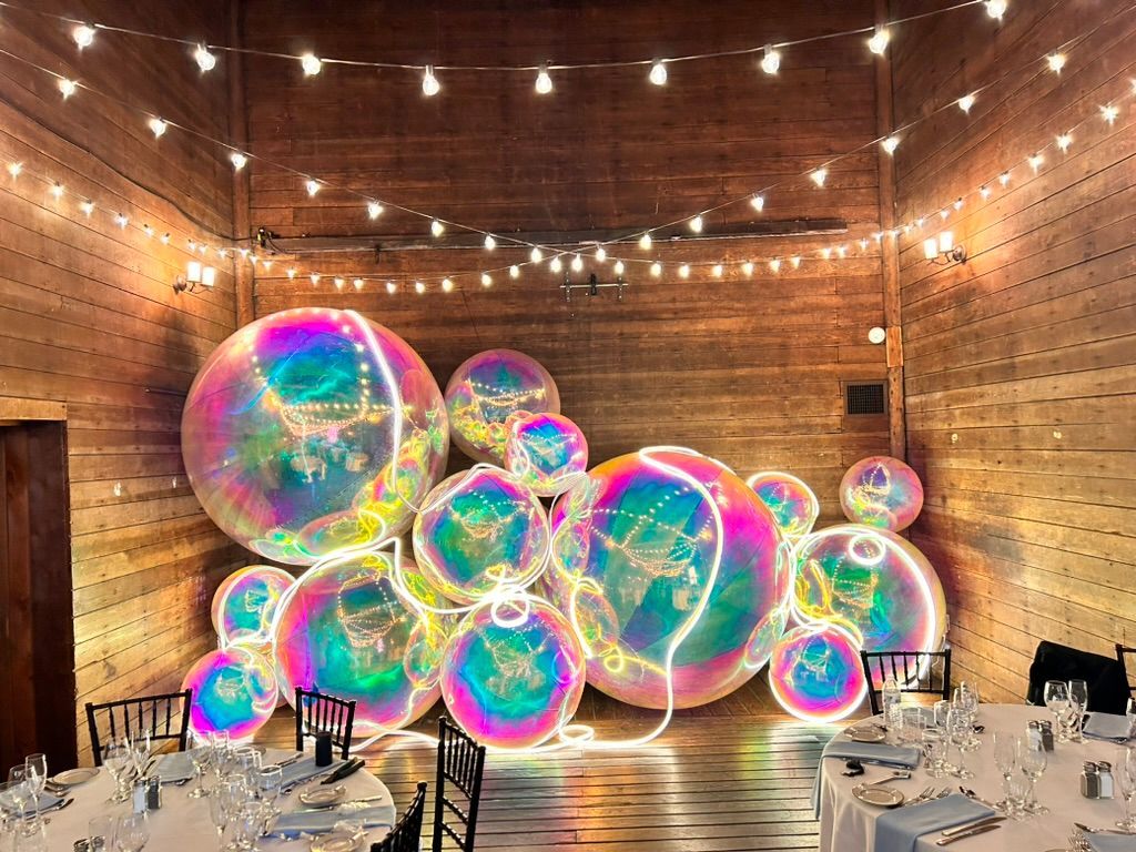Colorful giant soap bubbles in a wood-paneled event hall with string lights and tables set for dining