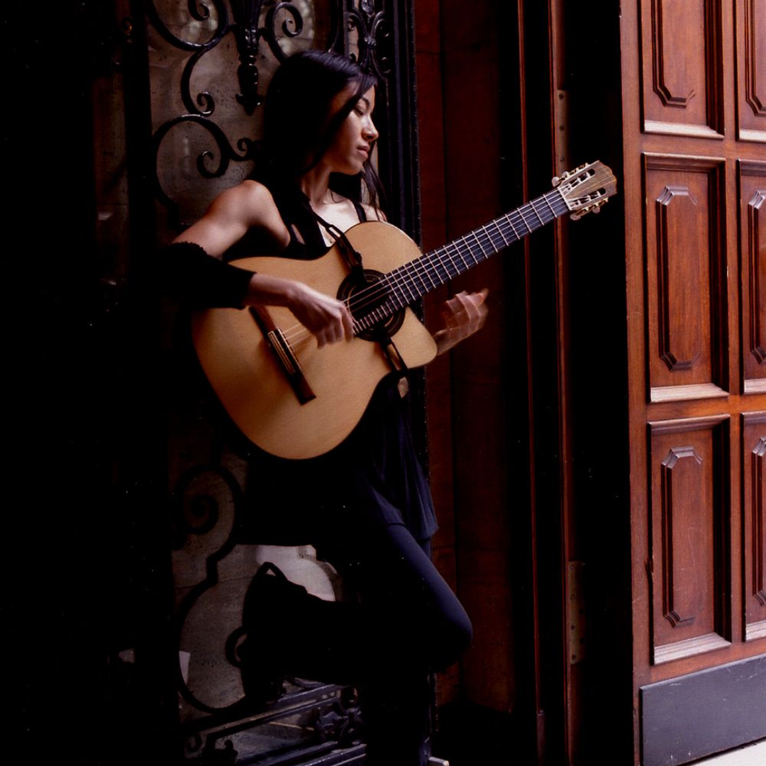Woman playing guitar in a dimly lit doorway beside ornate wooden doors