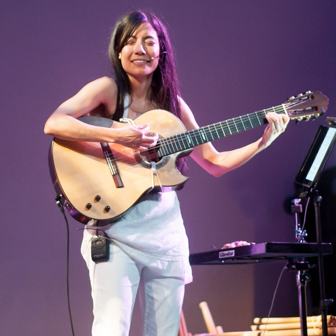 Musician smiling onstage, playing an acoustic guitar under purple lighting.