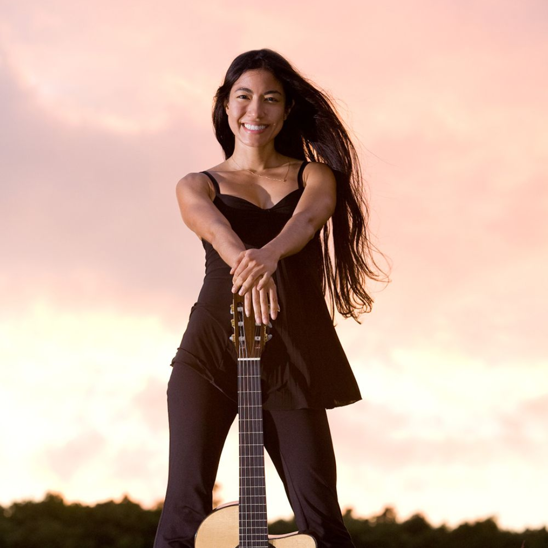 Woman standing with a guitar against a pink sunset sky