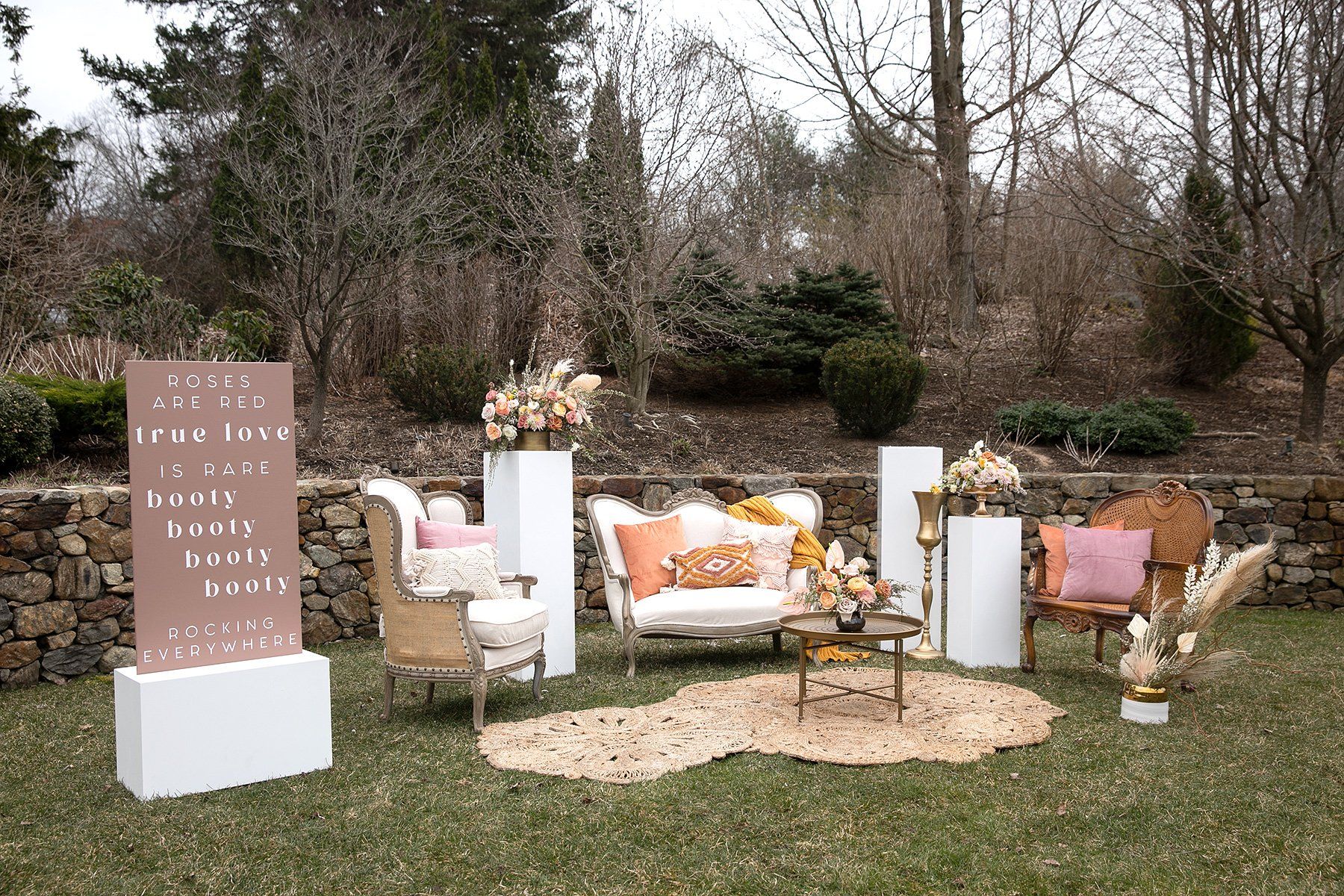 Outdoor wedding lounge with white furniture, peach accents, and a welcome sign in a garden setting