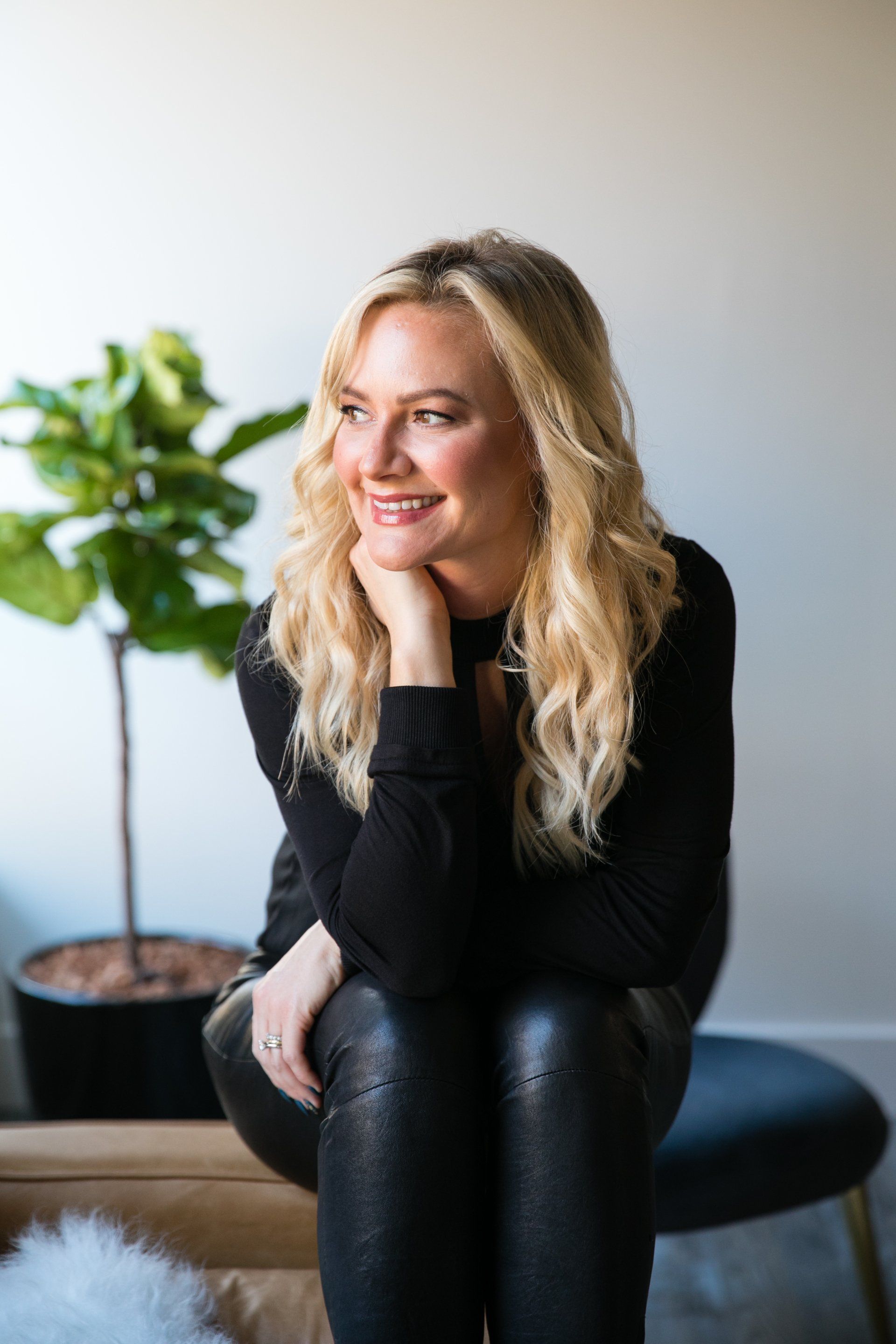 Smiling woman in black outfit seated on a bench beside a potted plant indoors