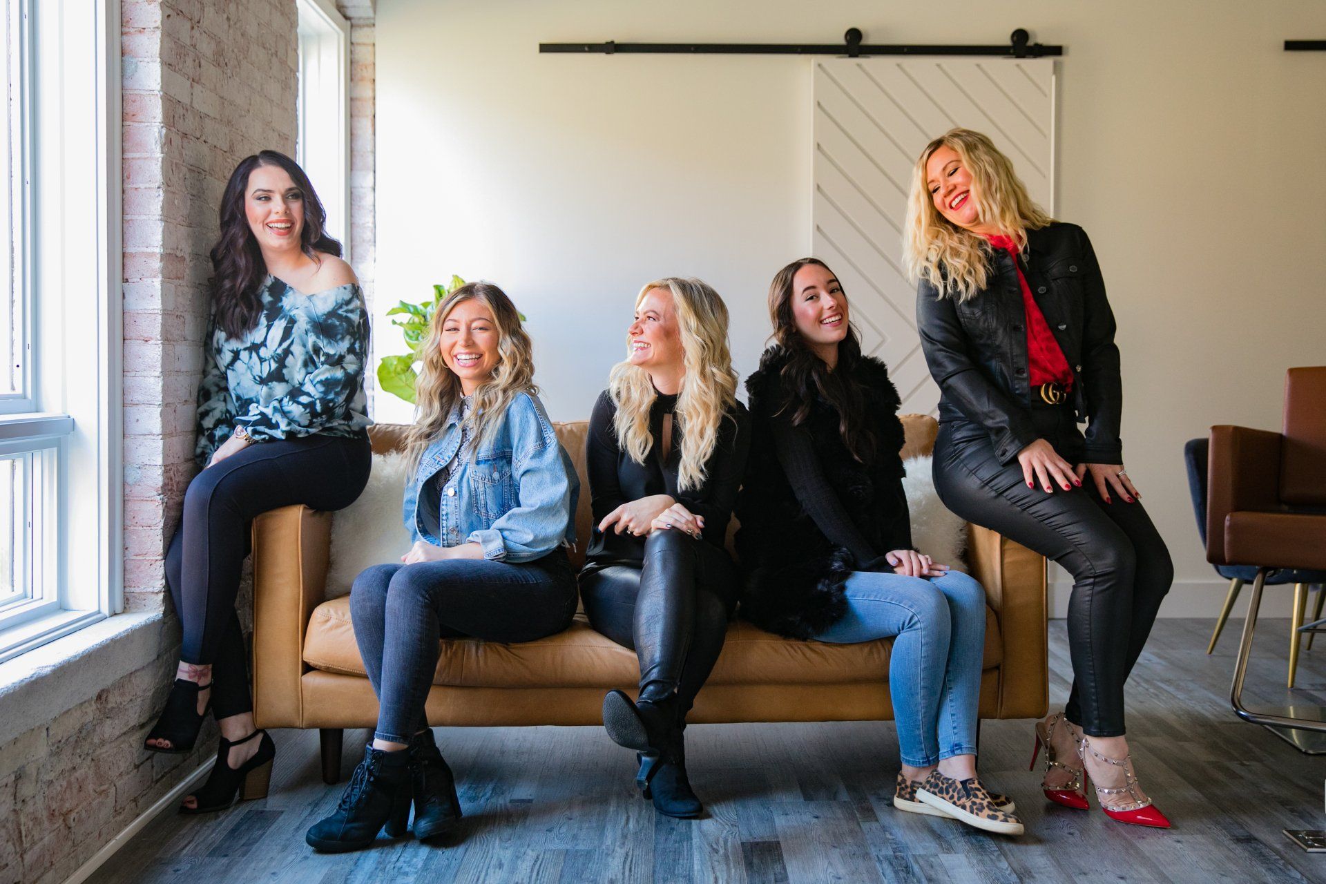Five women sit on a couch in a bright room, posing casually by a window and projection screen.