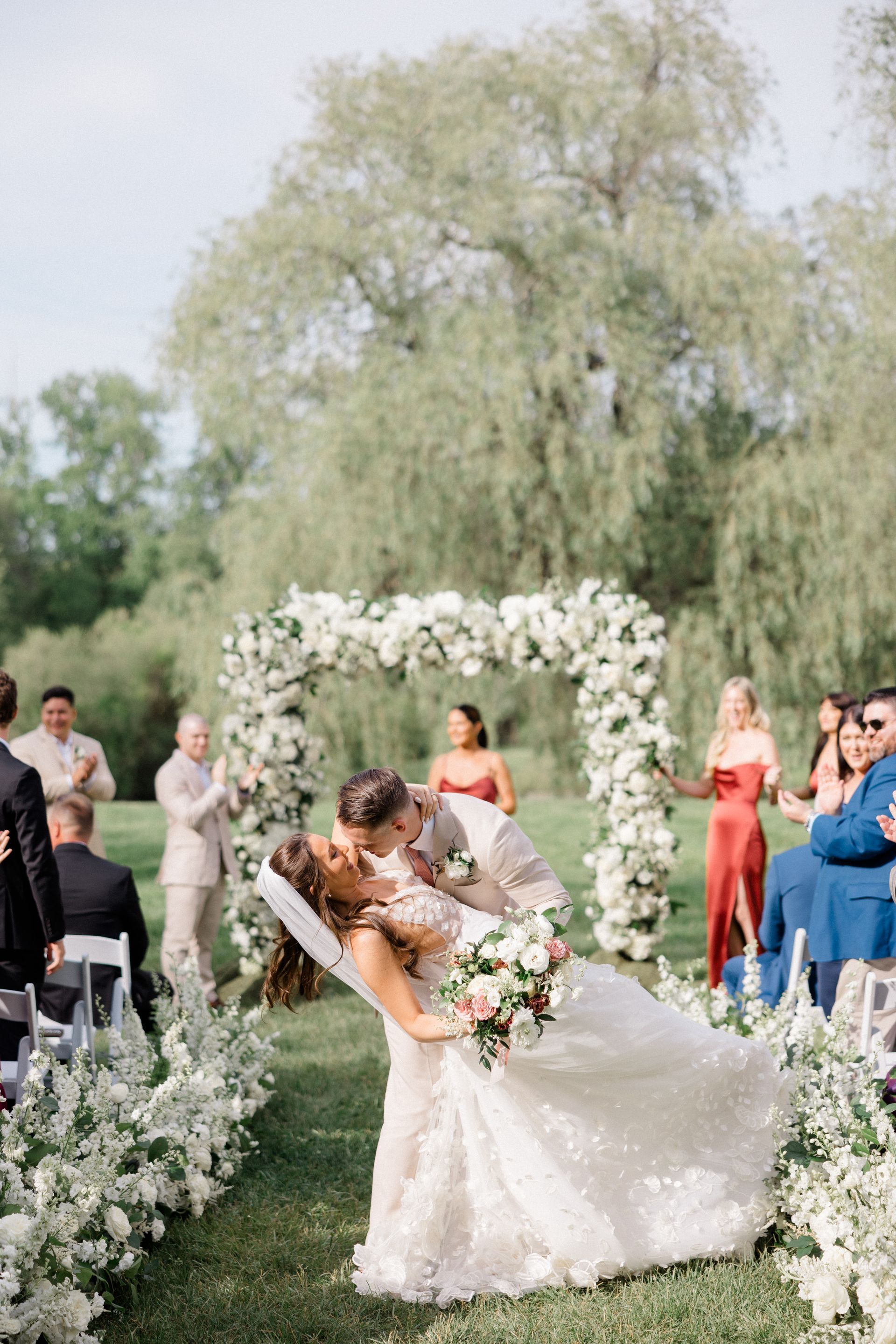Bride and groom kiss at outdoor wedding ceremony with white floral arch and guests watching
