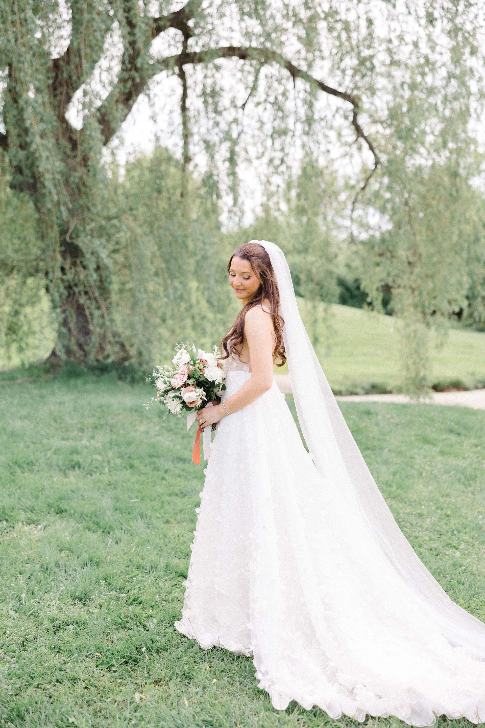 Bride in a white gown holding a bouquet, standing outdoors under a willow tree on a grassy lawn.