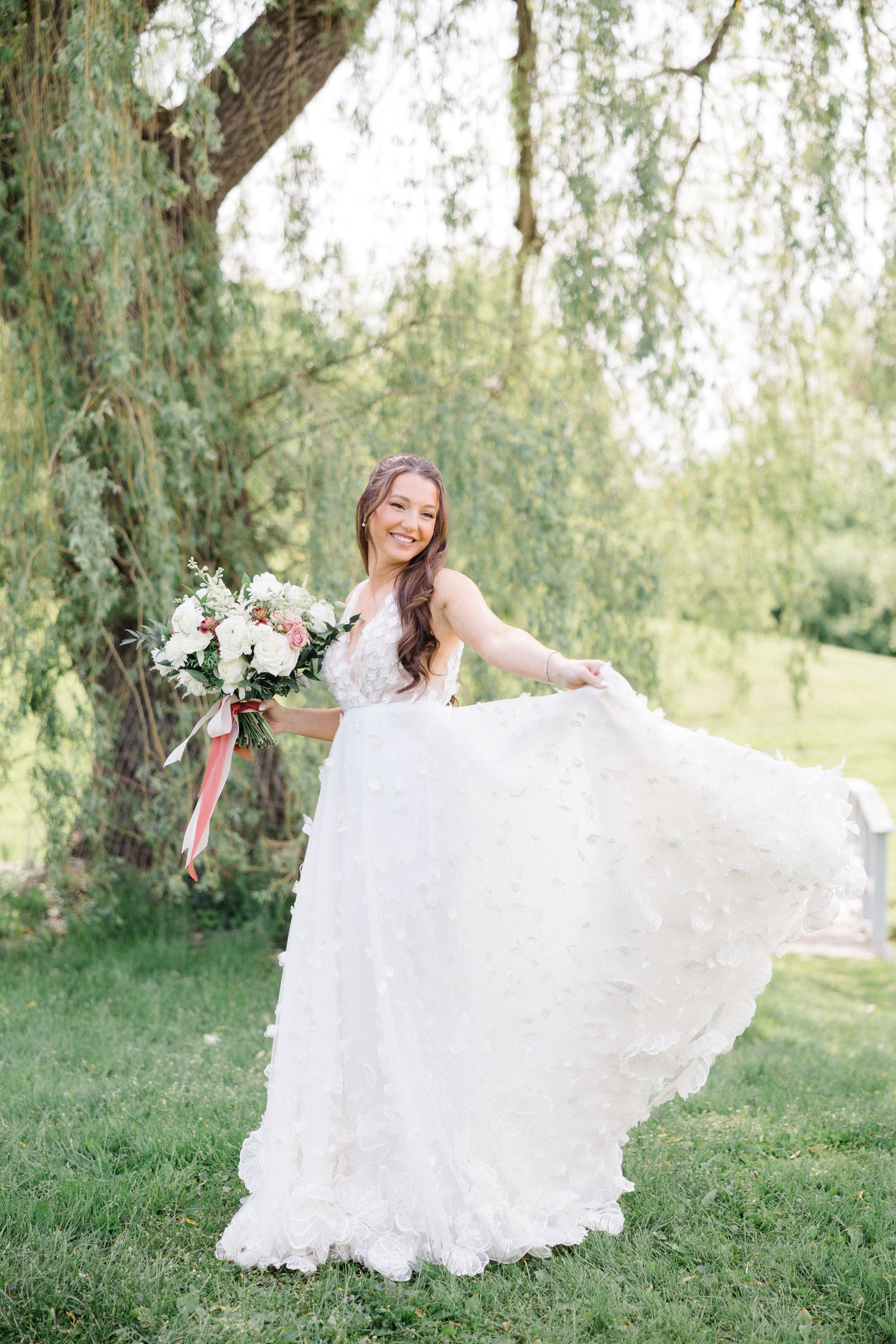 Bride in a white gown posing outdoors by a willow tree, holding a bouquet.