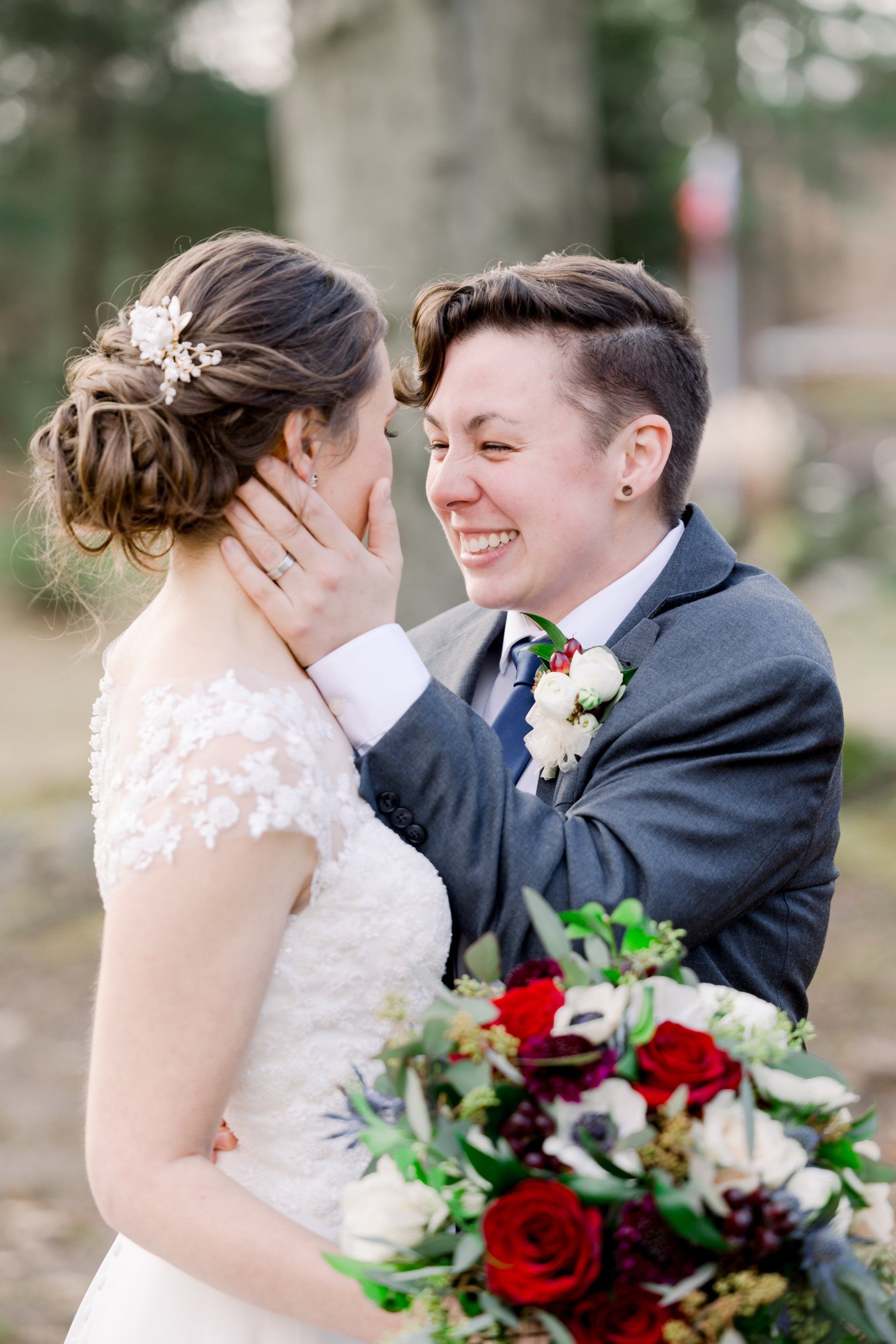 Two people in wedding attire smiling and touching faces, with a red-and-white bouquet in the foreground.