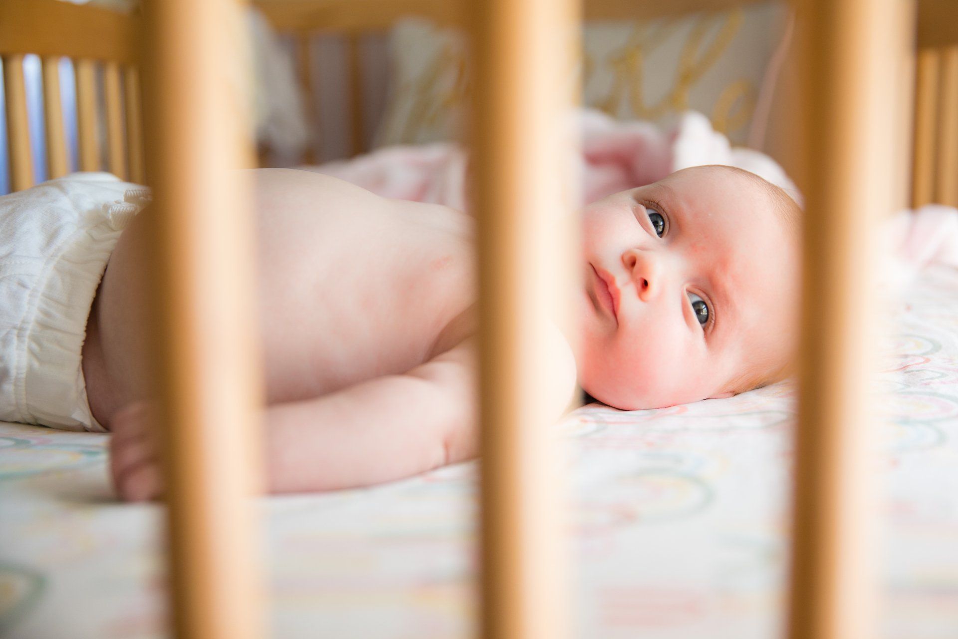 Baby lying in a crib, peeking through wooden slats with a calm expression