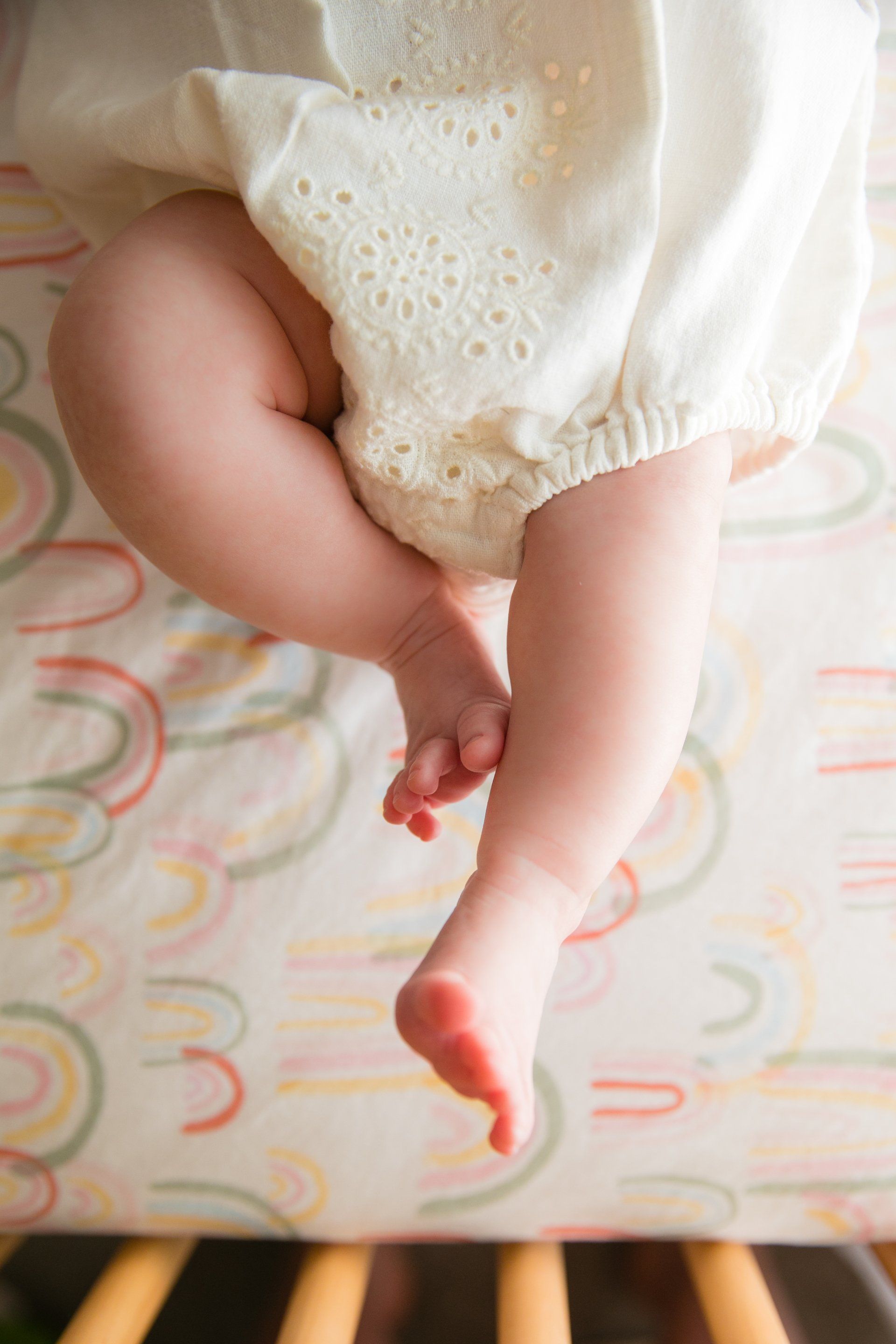 Baby's legs in a white outfit, lying on a patterned bedspread.