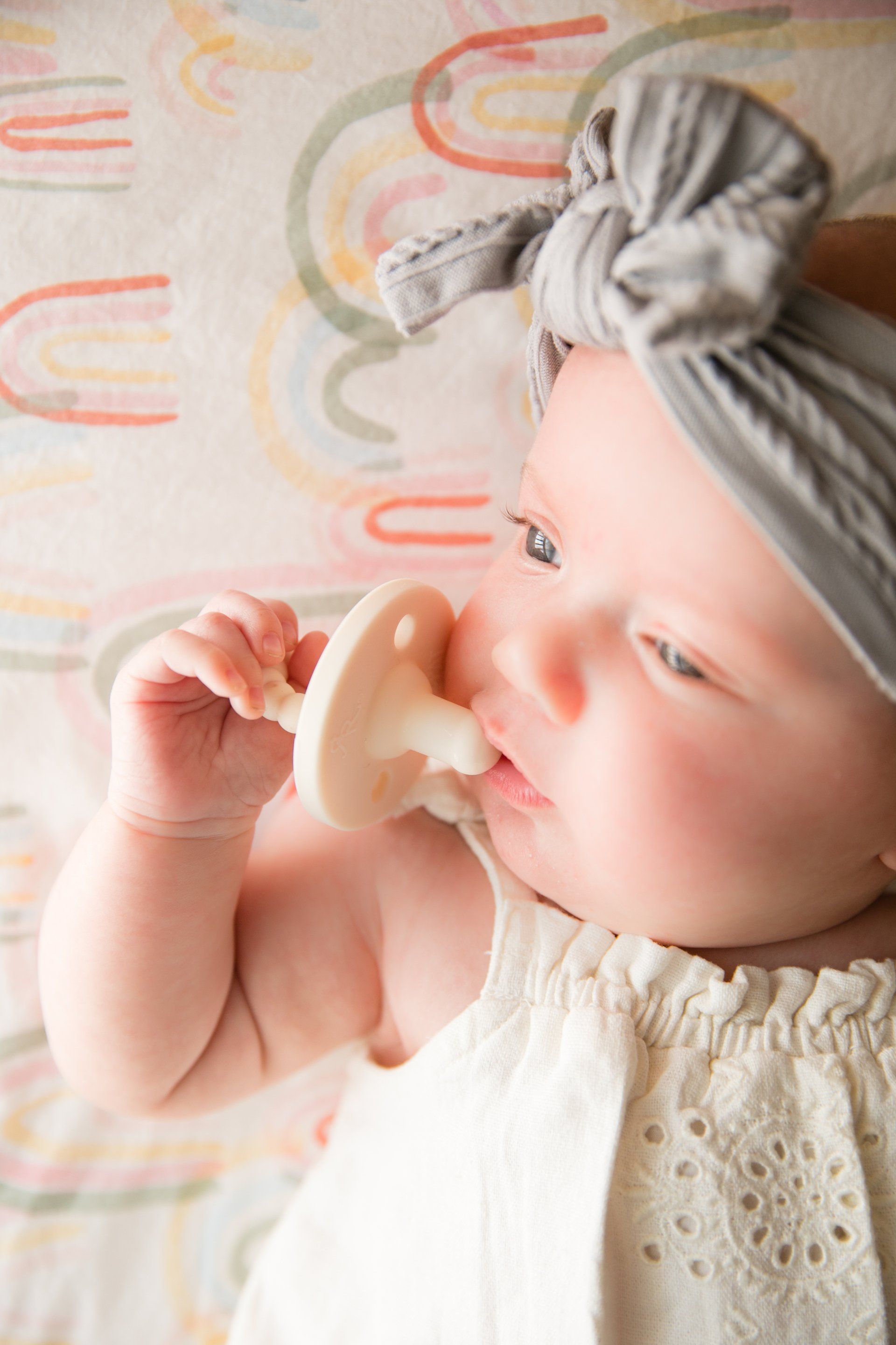 Baby in a gray headwrap holding a pacifier, resting against a patterned blanket.