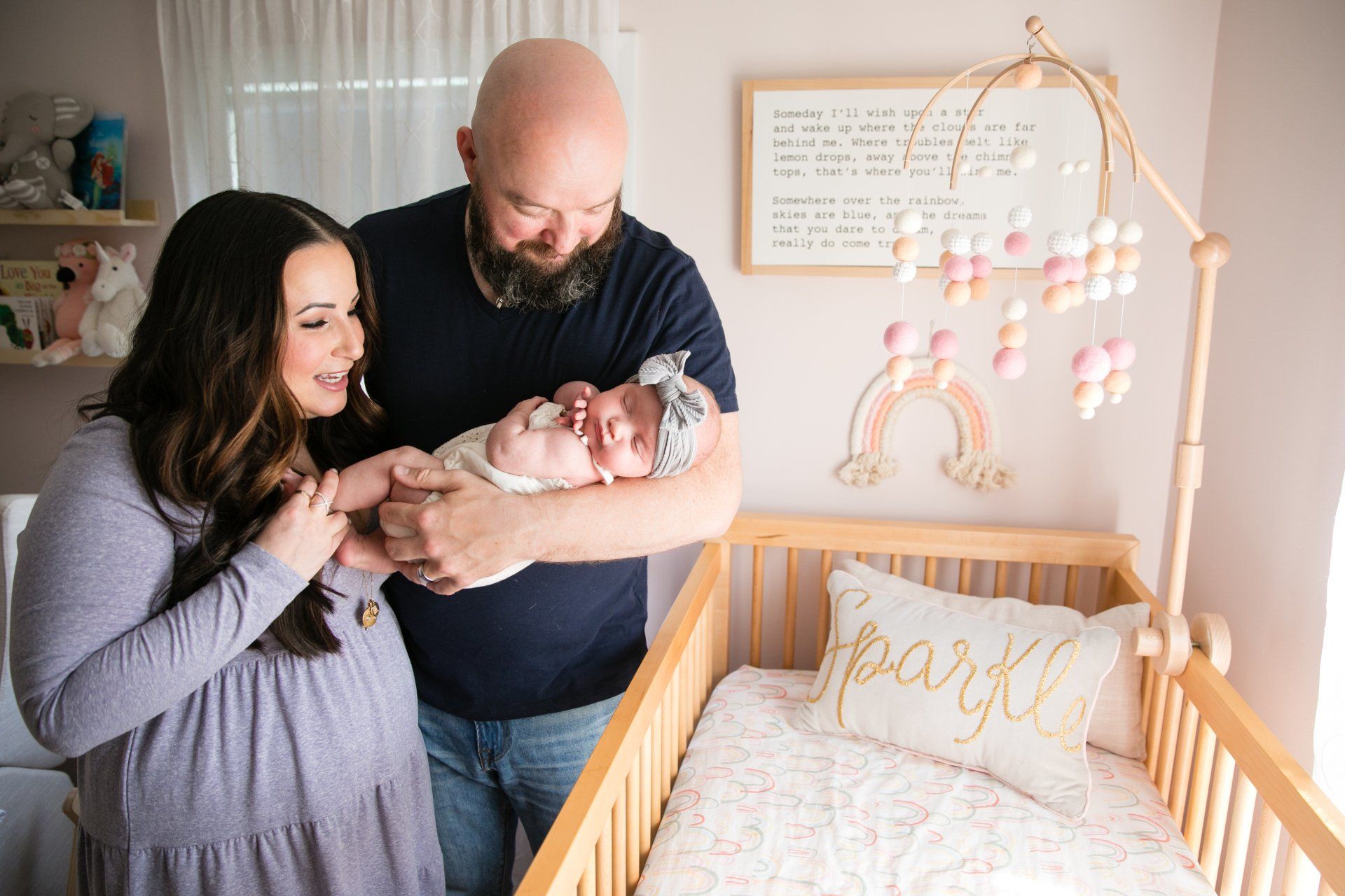 Parents holding newborn beside a crib with gold “hello” decor in a bright nursery