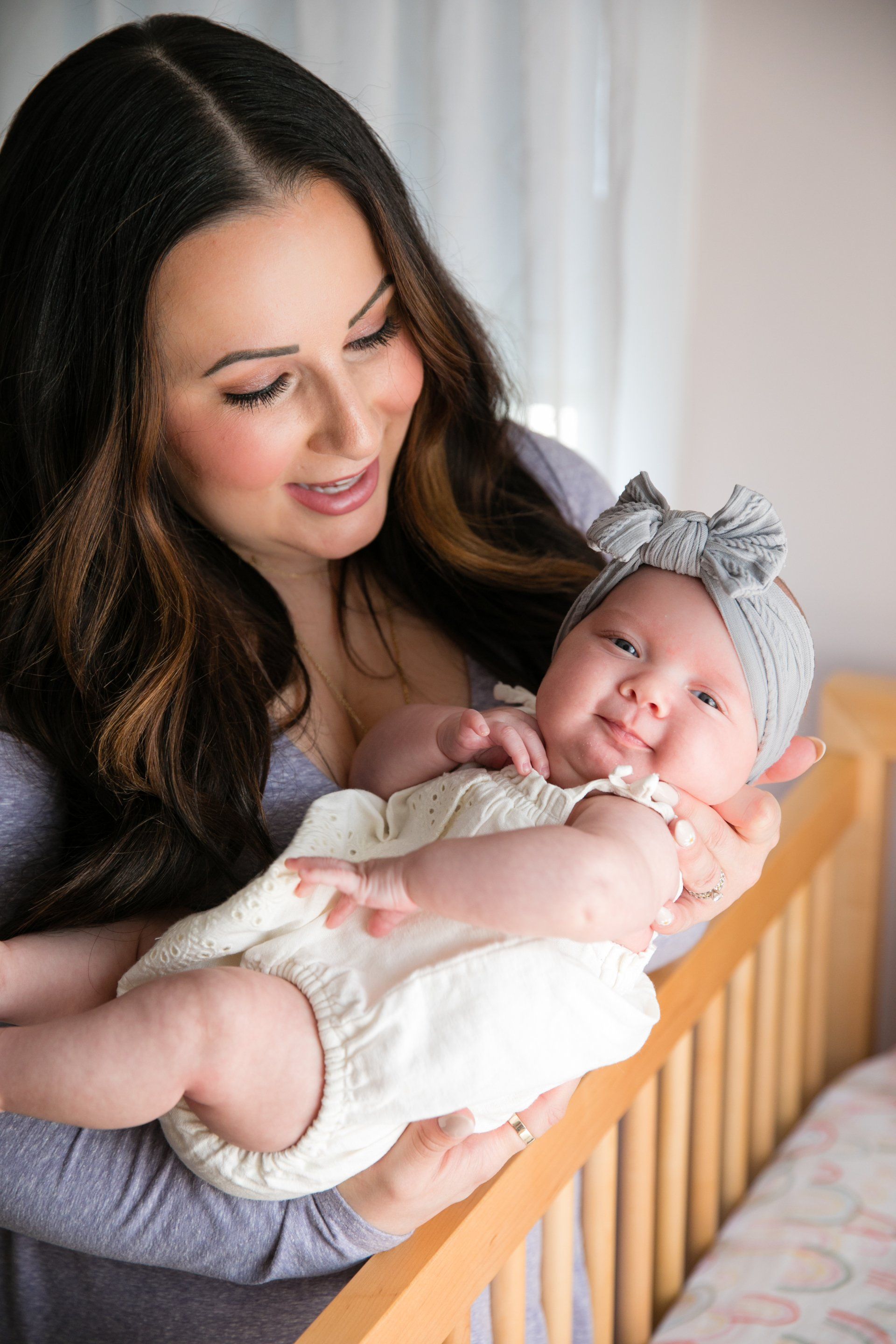 Woman smiling and holding a sleeping baby in a wooden crib, both in soft daylight.