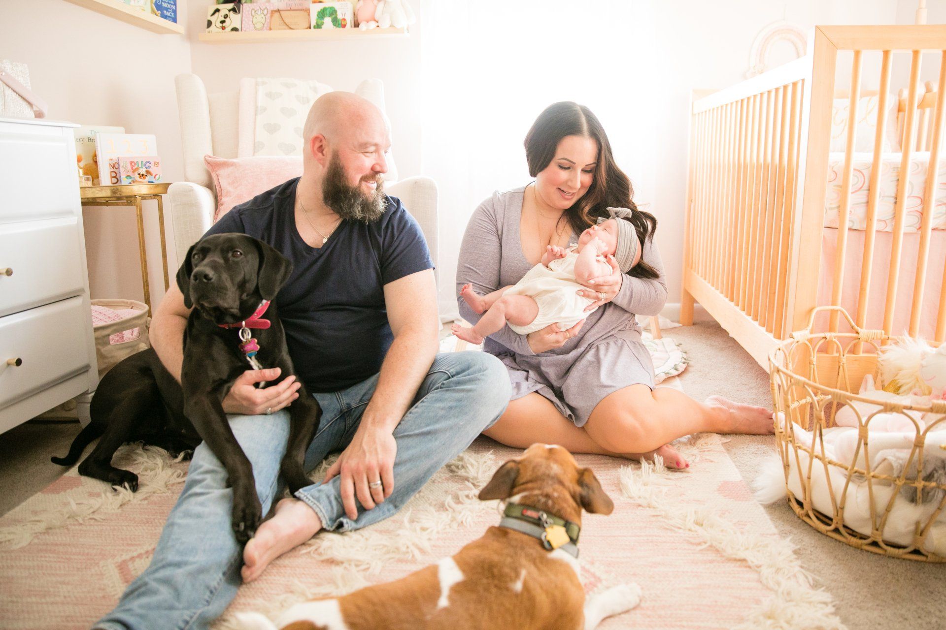 Couple sitting on a nursery floor with two dogs and a baby in a crib nearby