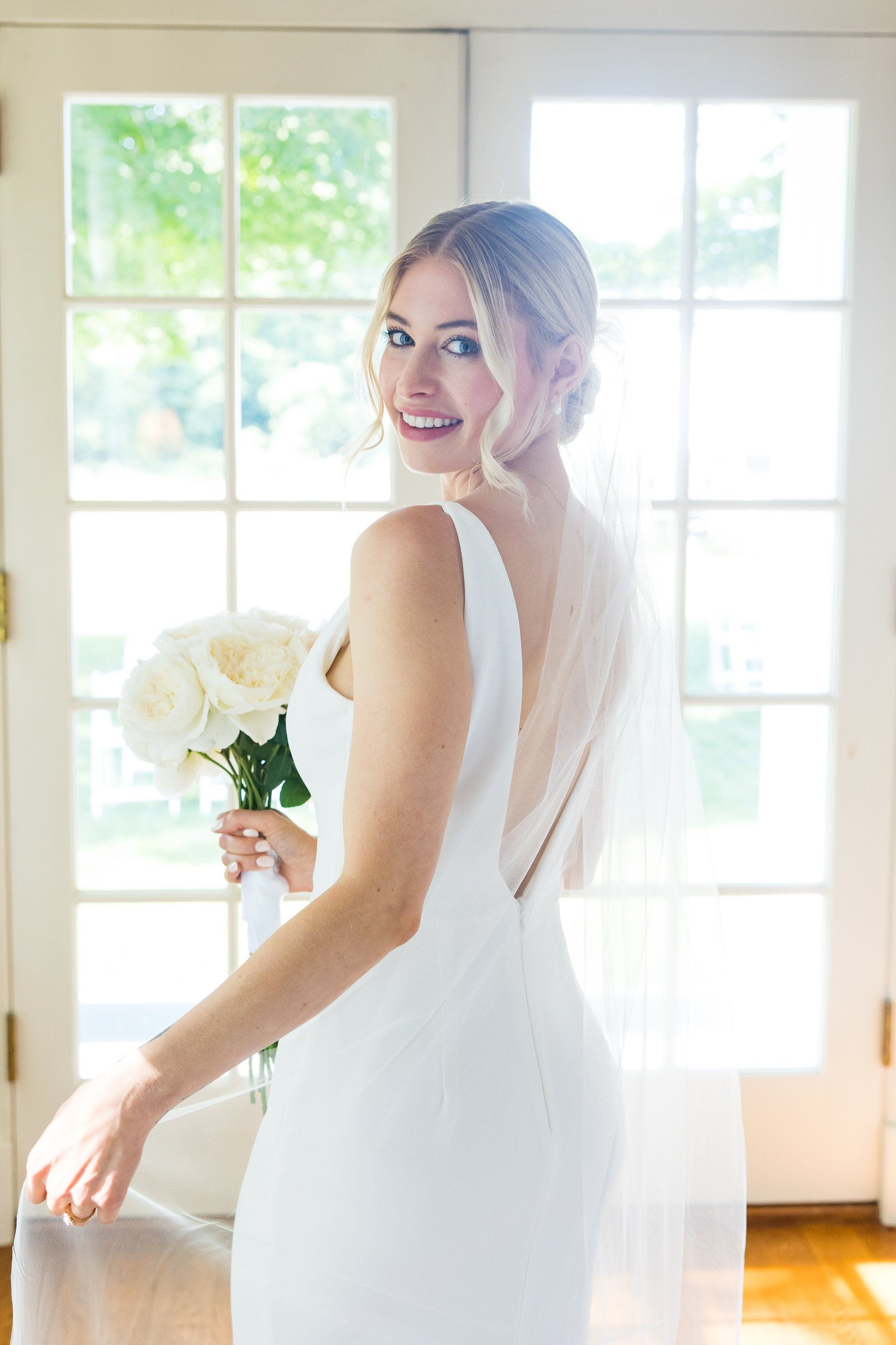 Bride in a white gown holding a bouquet, turning back and smiling by bright windows.