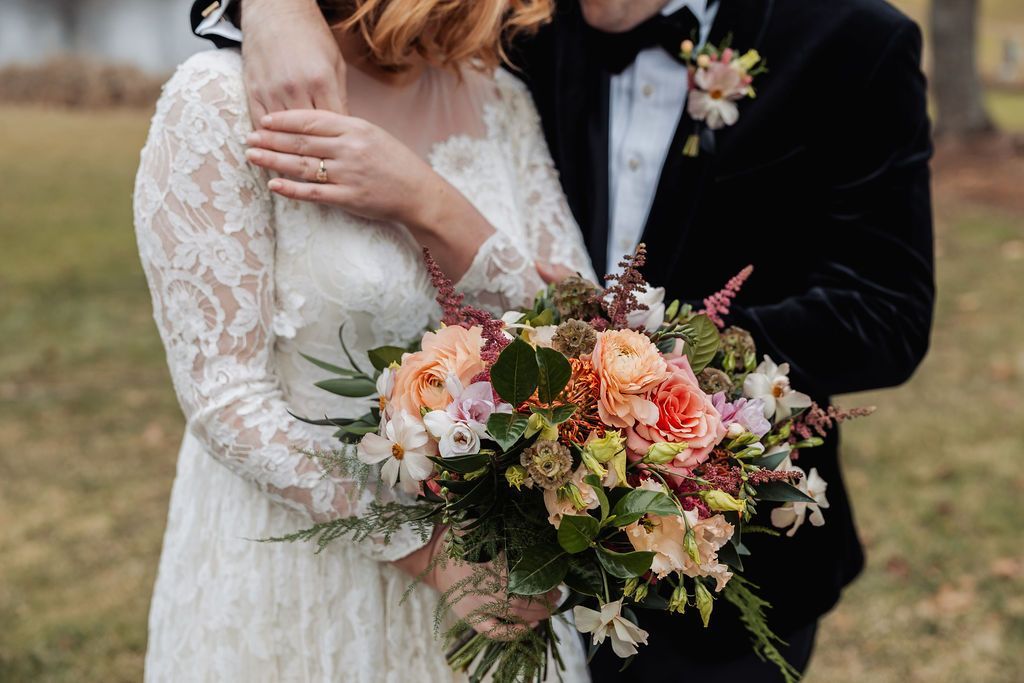 Wedding couple holding a colorful bouquet, with the bride in a lace dress and groom in a black suit outdoors