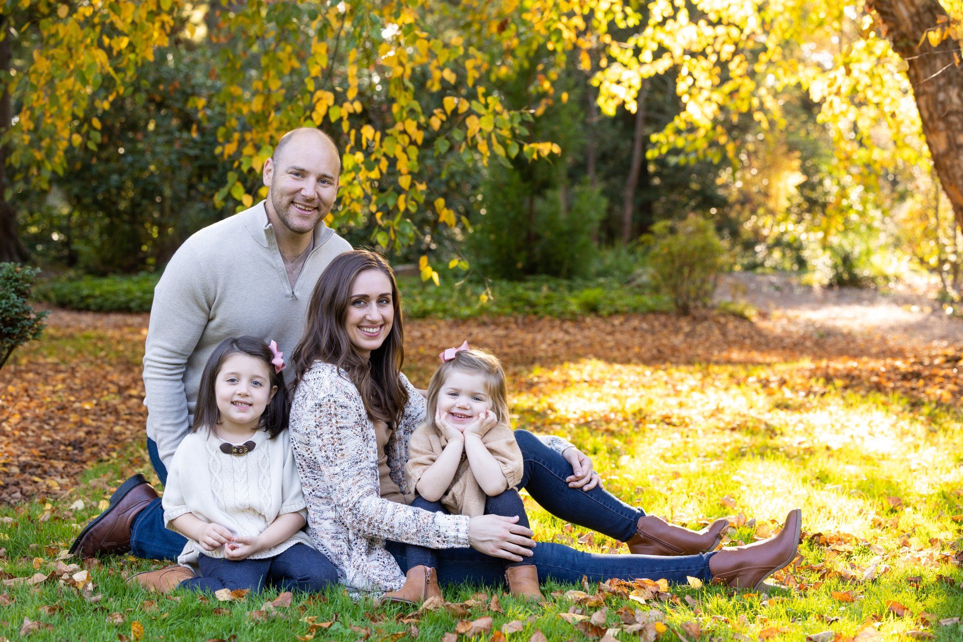 Family of four sitting on grass in a sunlit autumn park, smiling at the camera