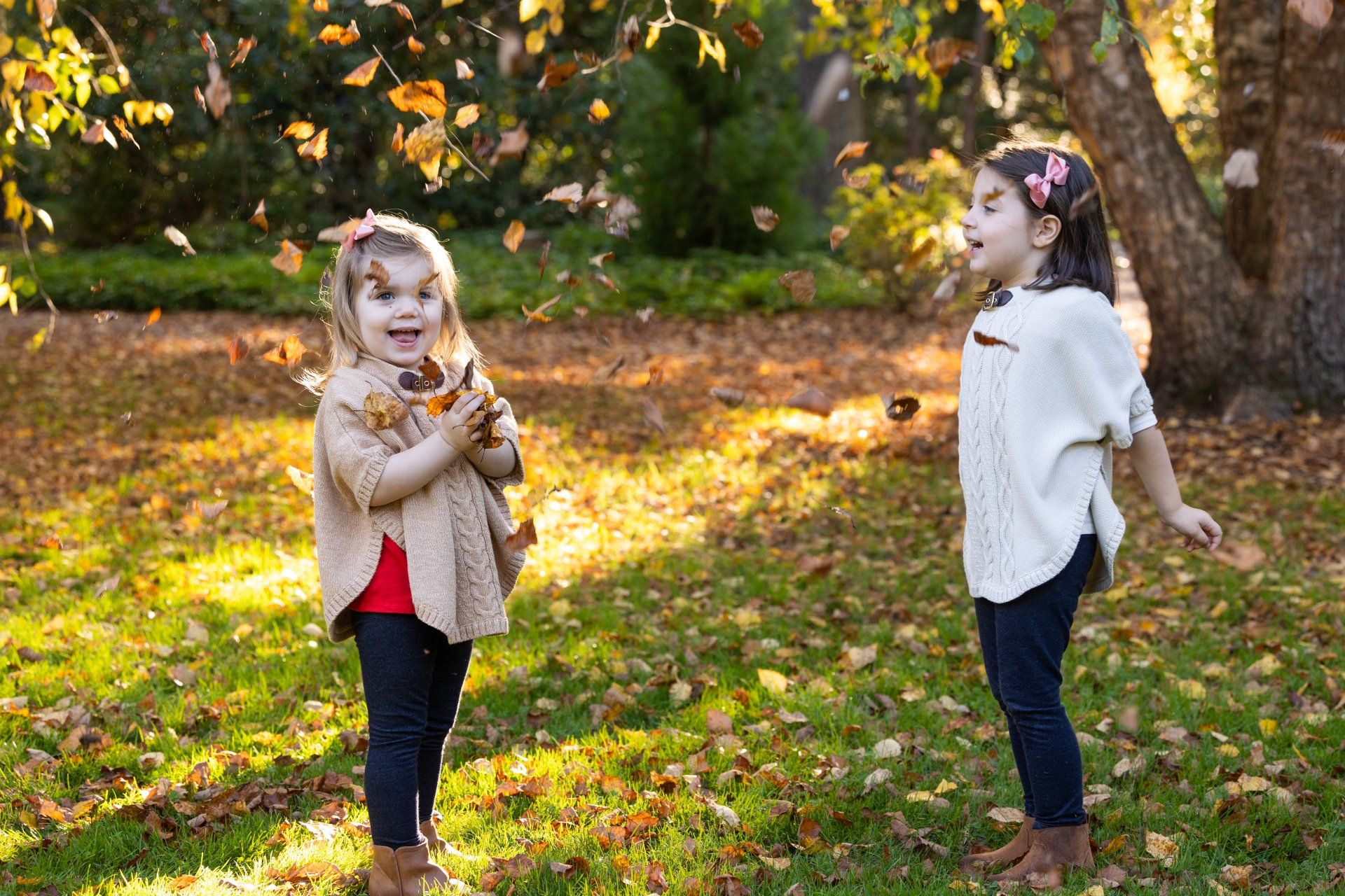 Two children standing in a sunny autumn park, one holding leaves and the other posing by a tree.