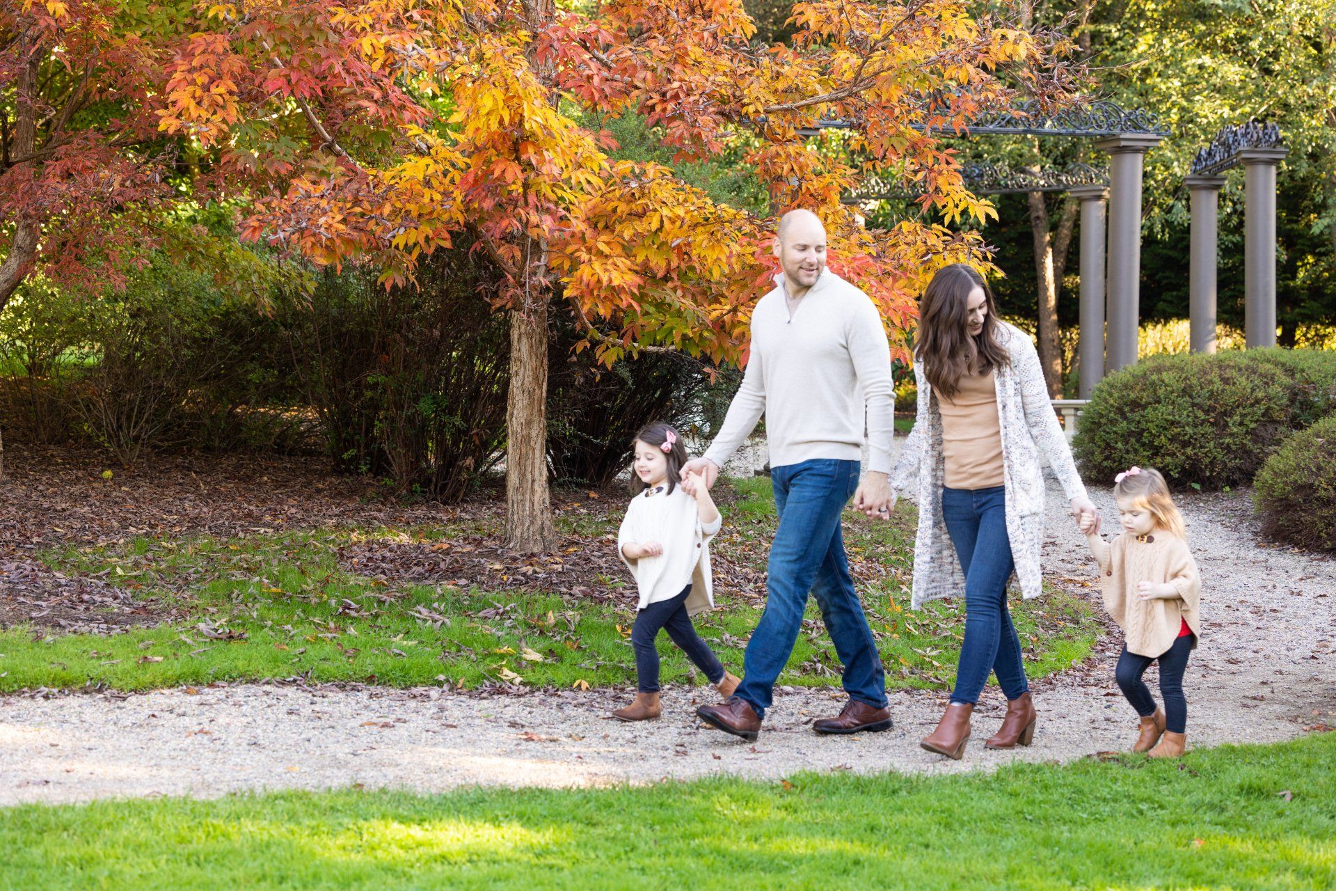 Family walking on a garden path beside autumn trees and a pergola