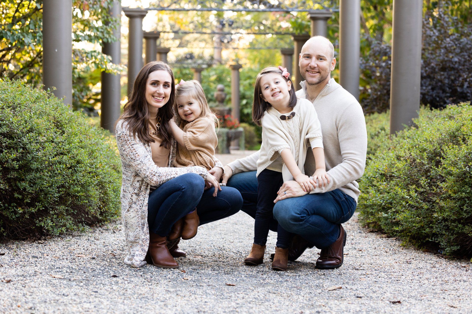 Family crouching together in a garden path, smiling among shrubs and wooden posts