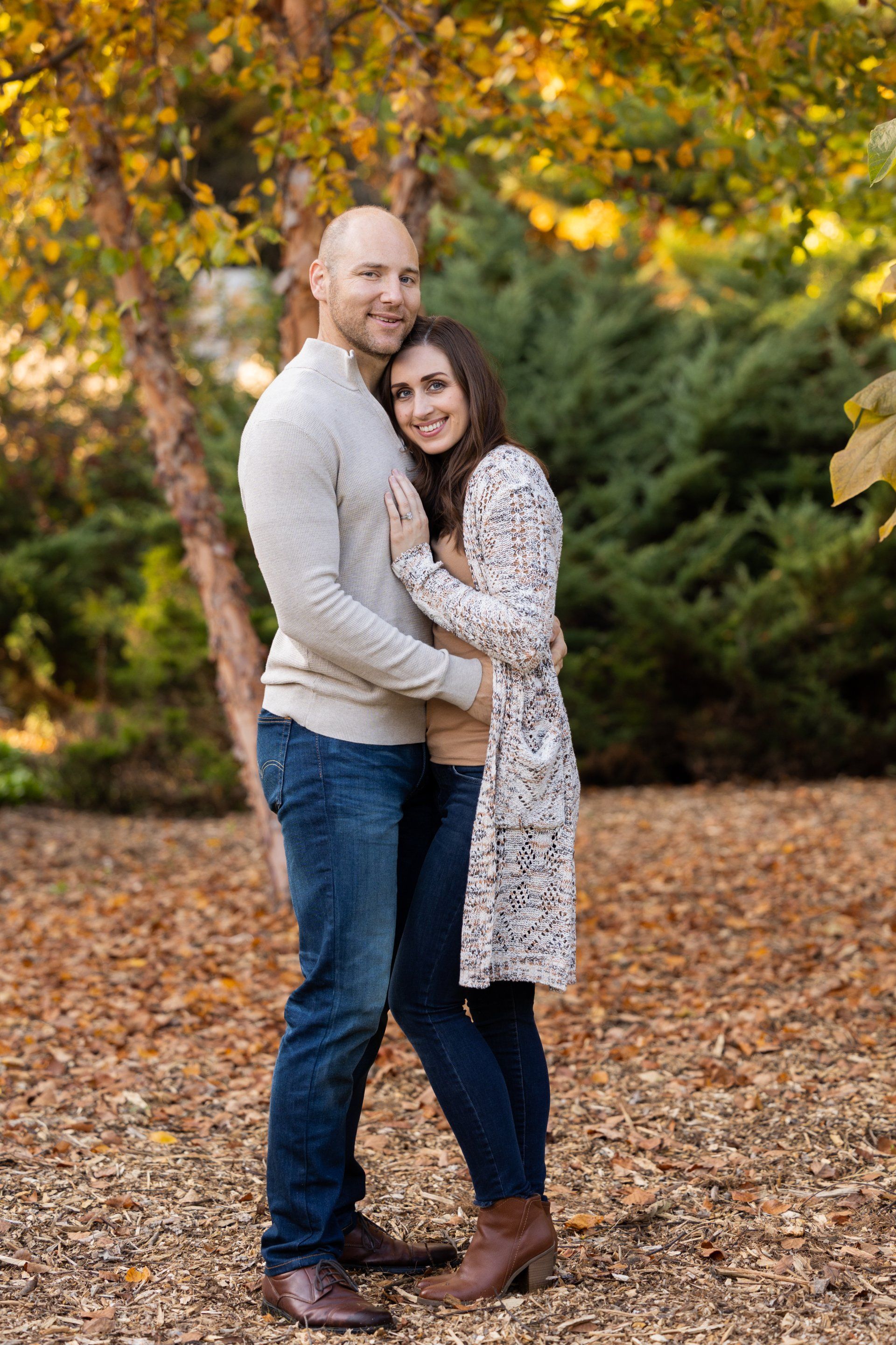 Couple hugging outdoors in a leafy autumn park, smiling in sweaters and jeans.