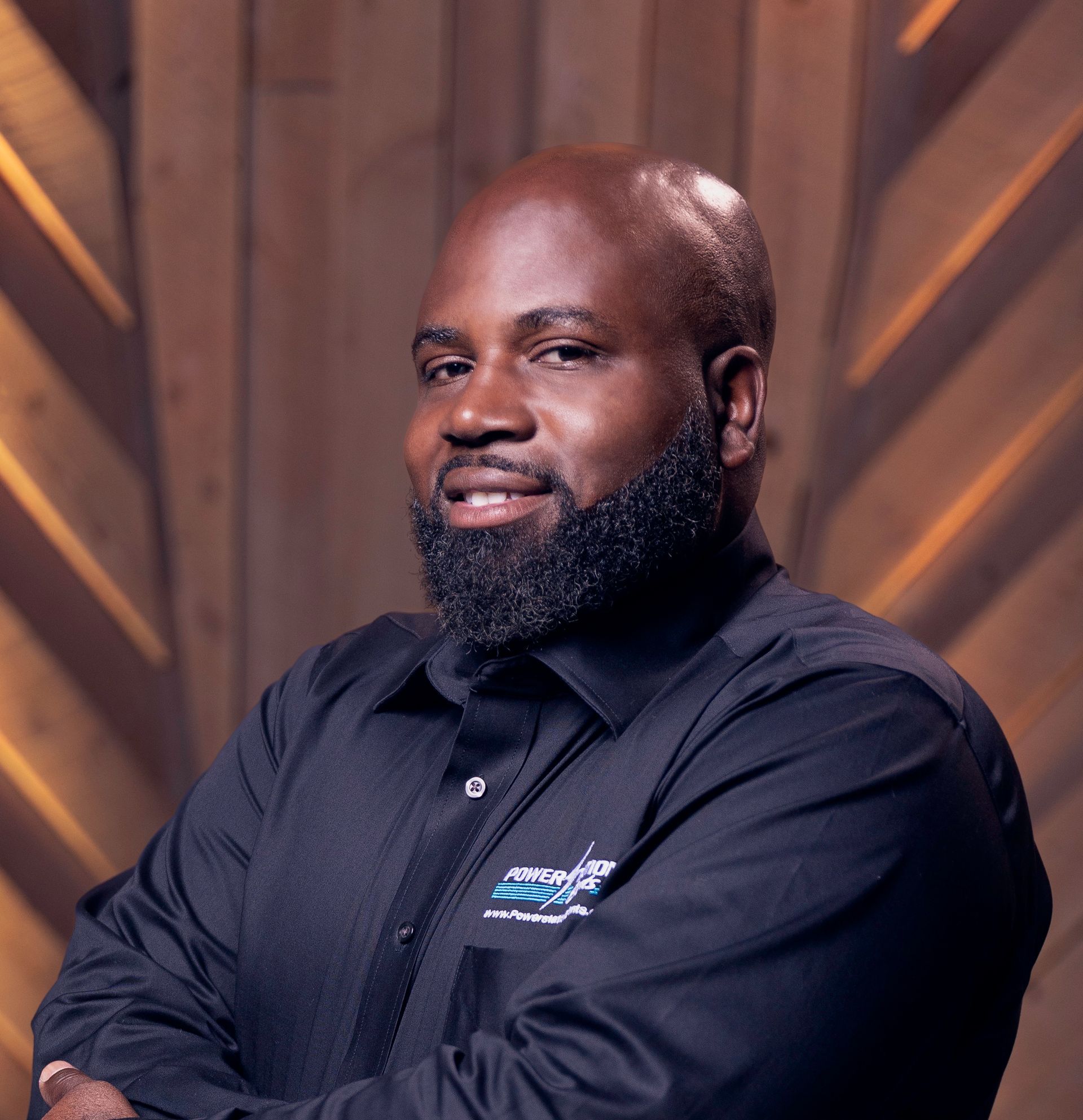 Portrait of a smiling man in a black shirt with arms crossed against a wooden backdrop