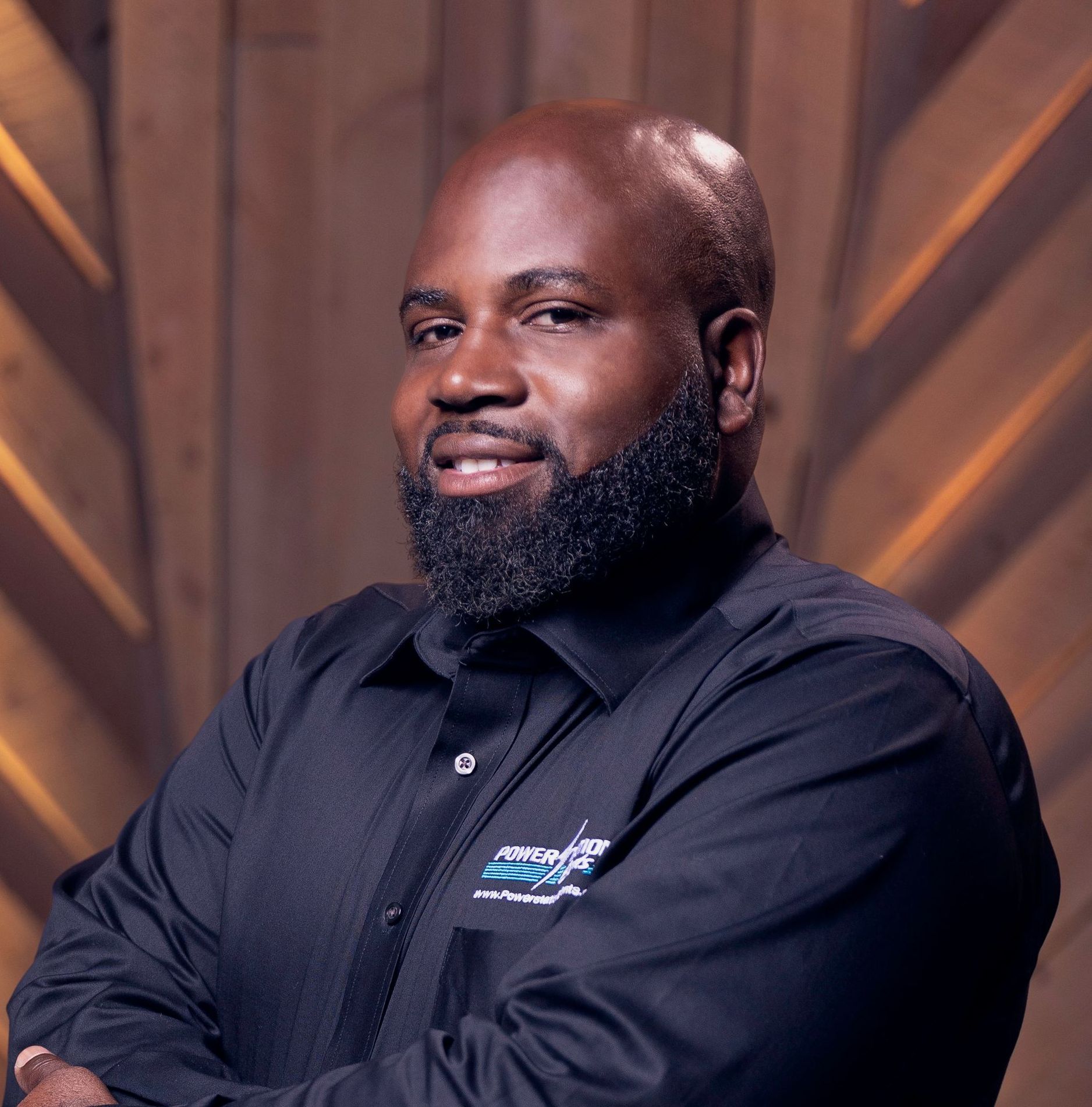 Smiling man in a black shirt with arms crossed against a warm wooden background