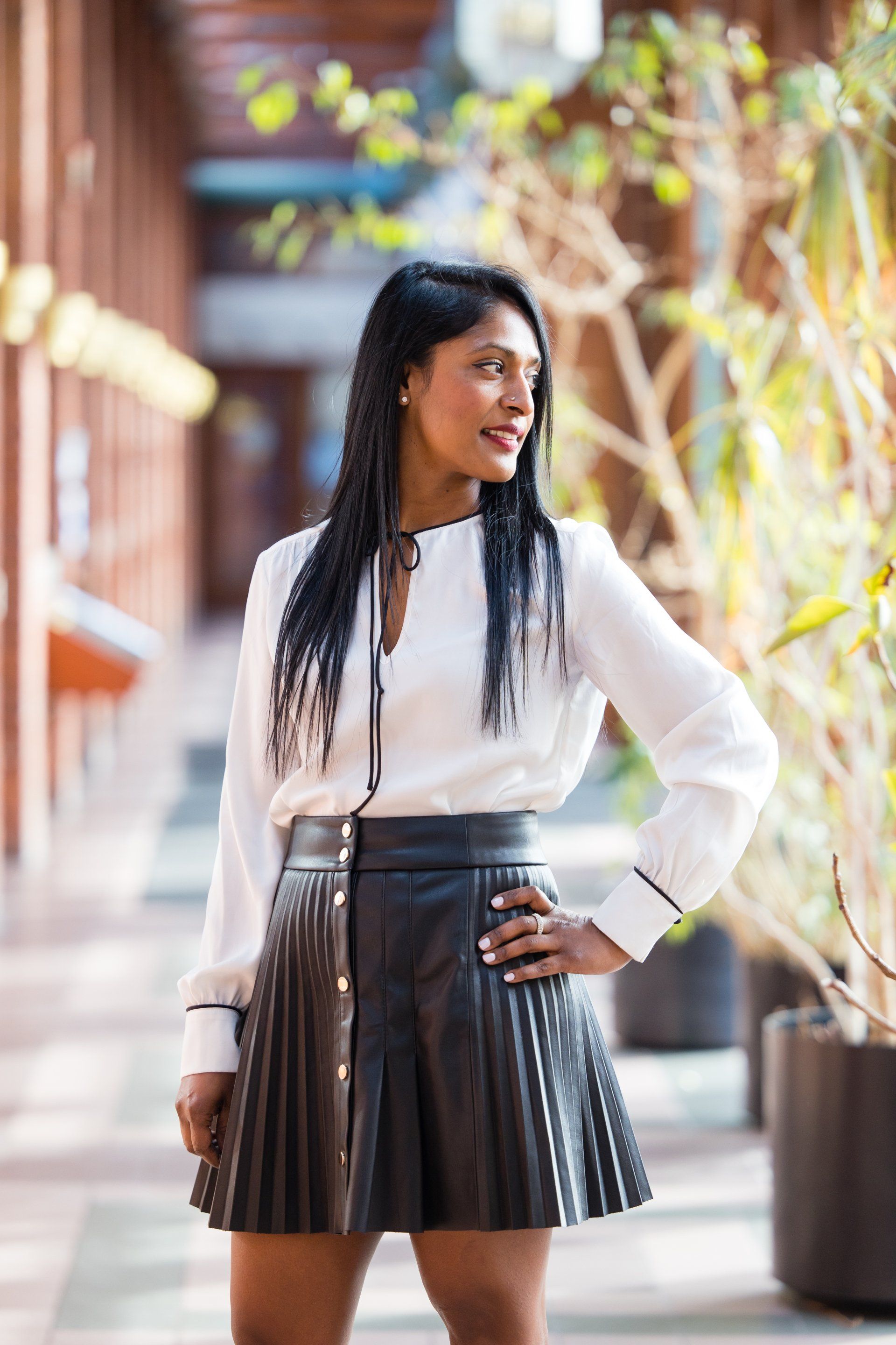 Woman in white blouse and black pleated skirt standing outdoors on a sunlit walkway.