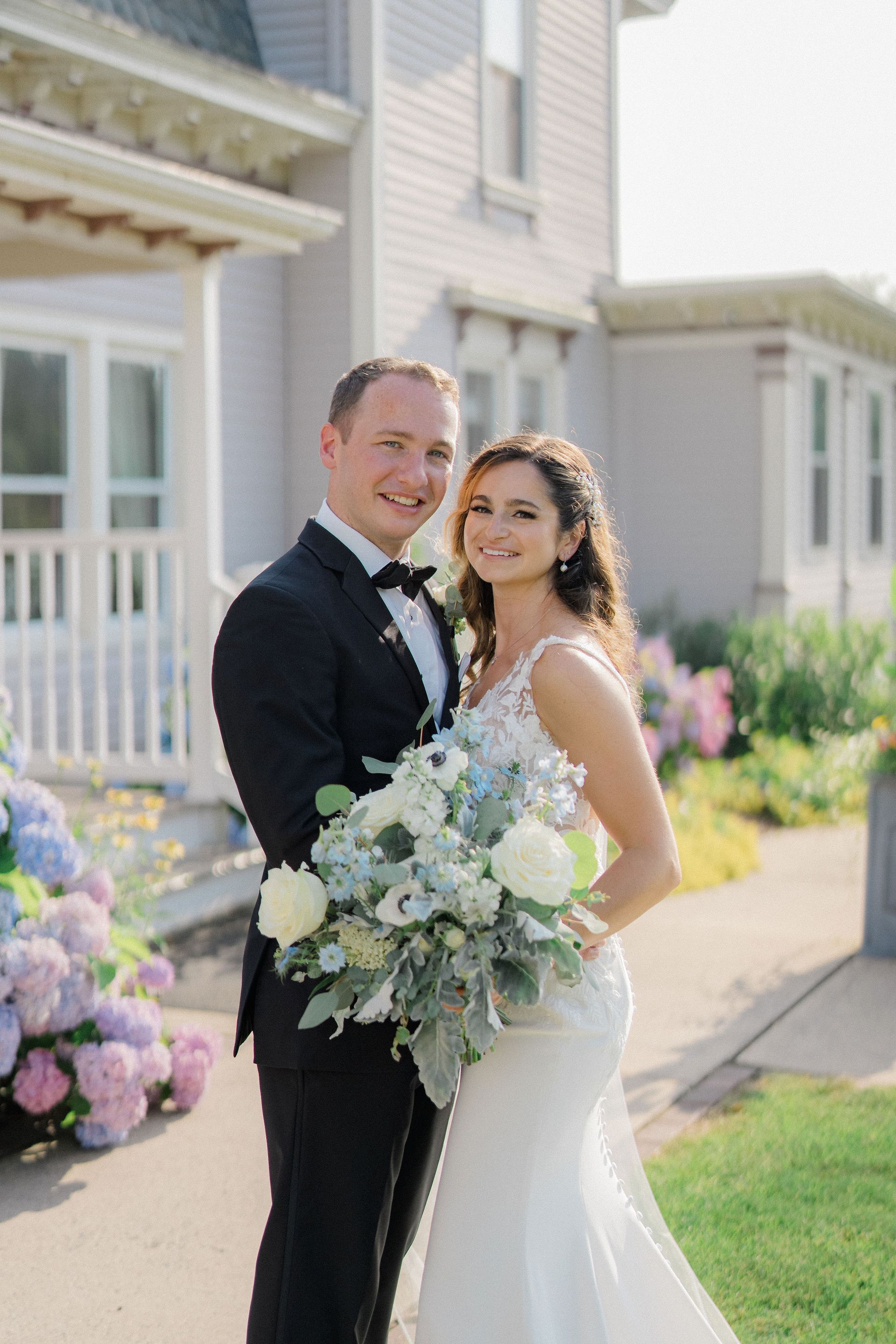 Newlywed couple posing outside a house, holding a white floral bouquet, smiling in formal wedding attire