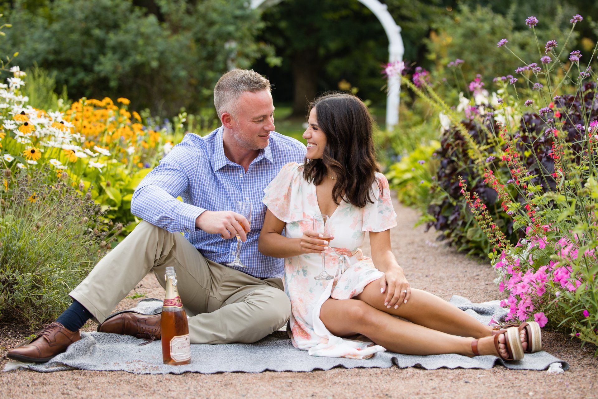 Couple sitting on a garden path, smiling and sharing a drink among colorful flowers.