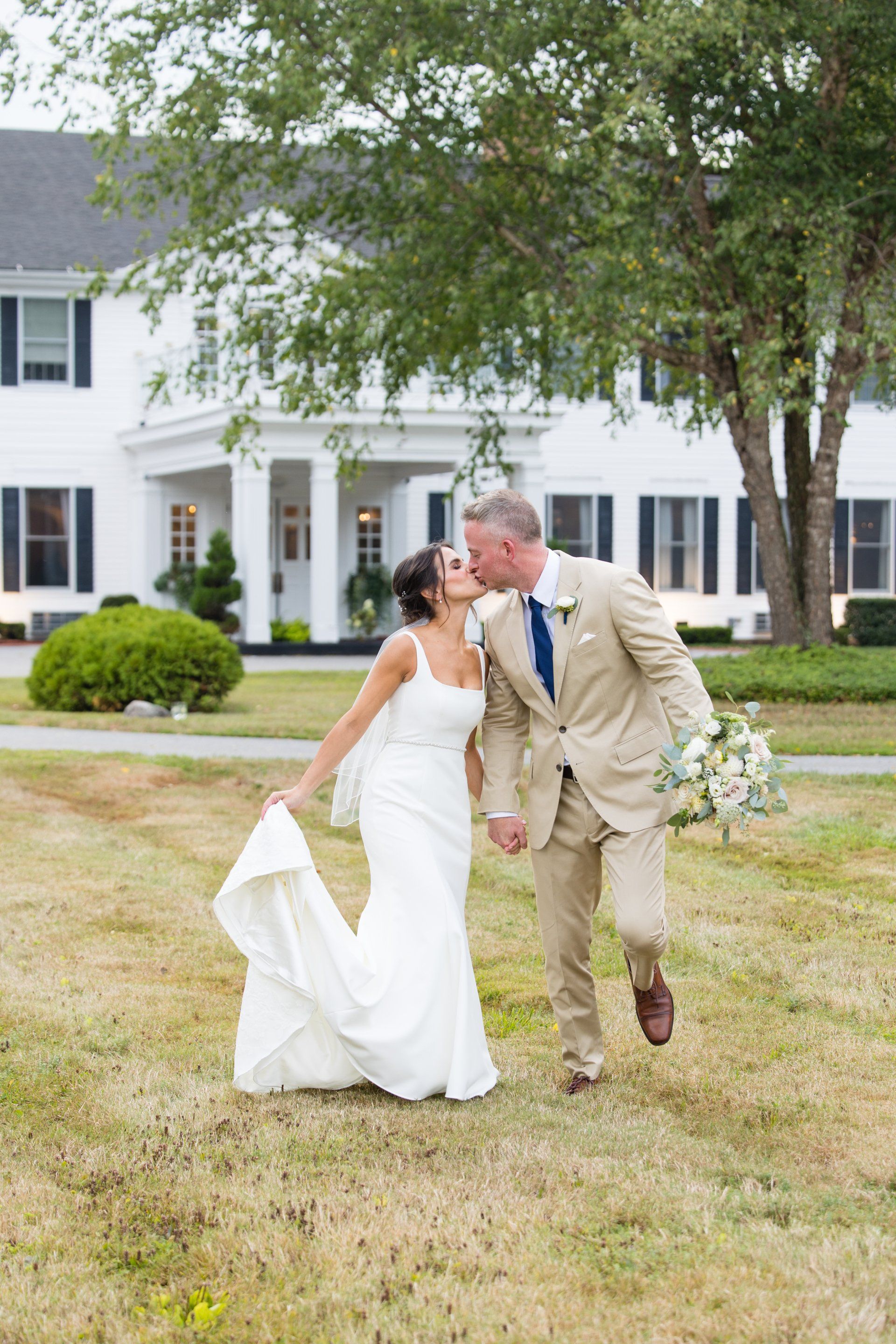 Bride and groom in wedding attire kiss on a lawn in front of a white house.