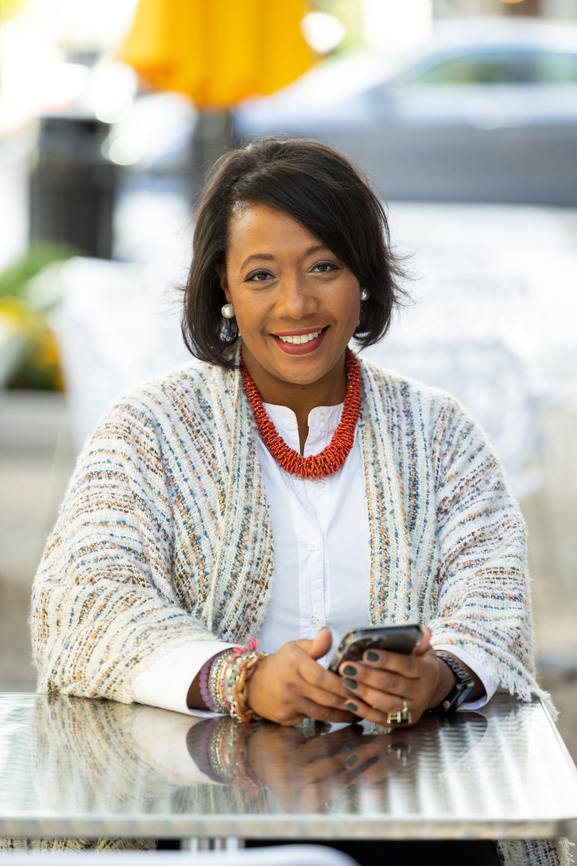 Smiling woman in white sweater and red necklace, seated at a table outdoors holding a phone