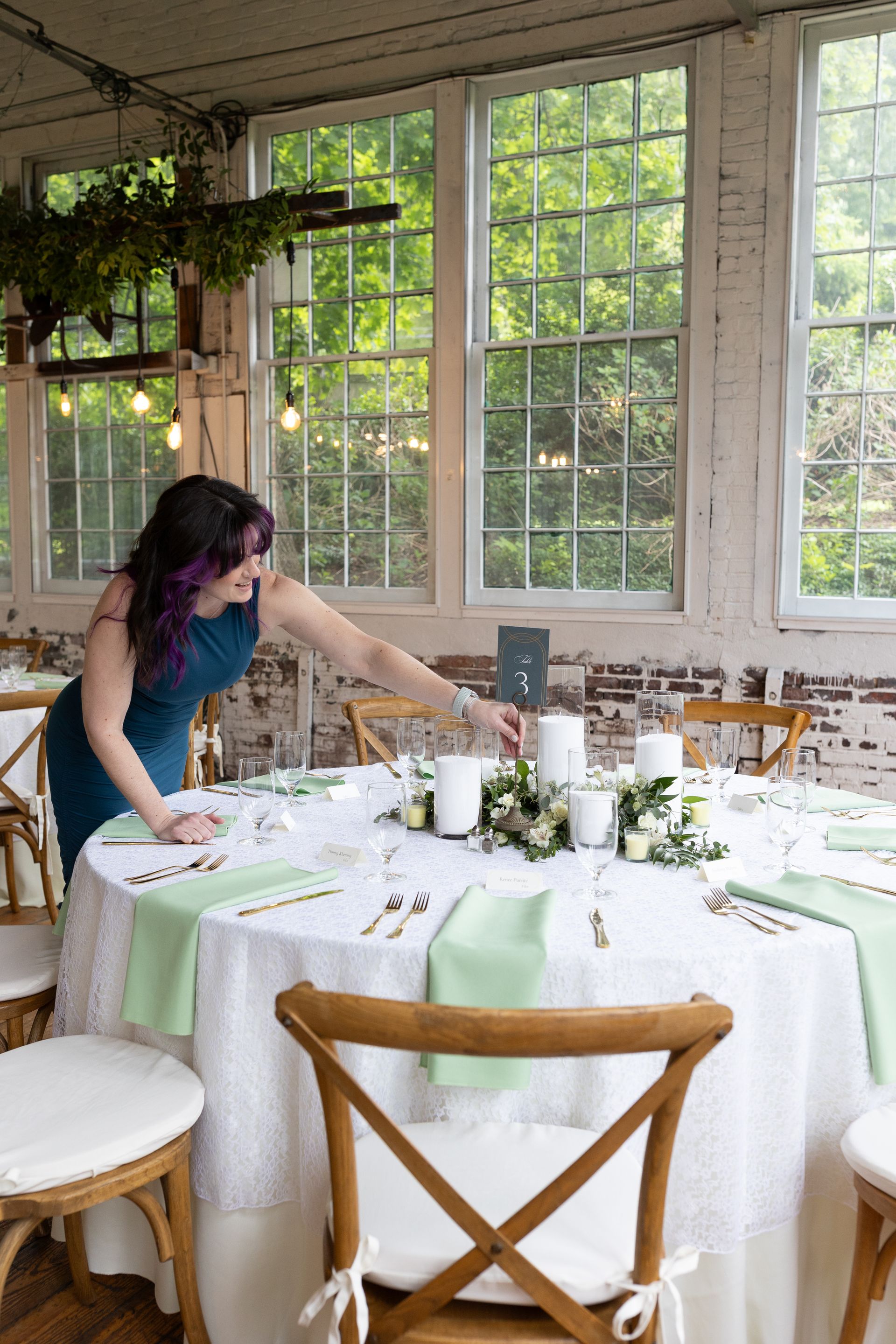 Woman arranging a round dining table with white linens and green accents in a sunlit room with large windows