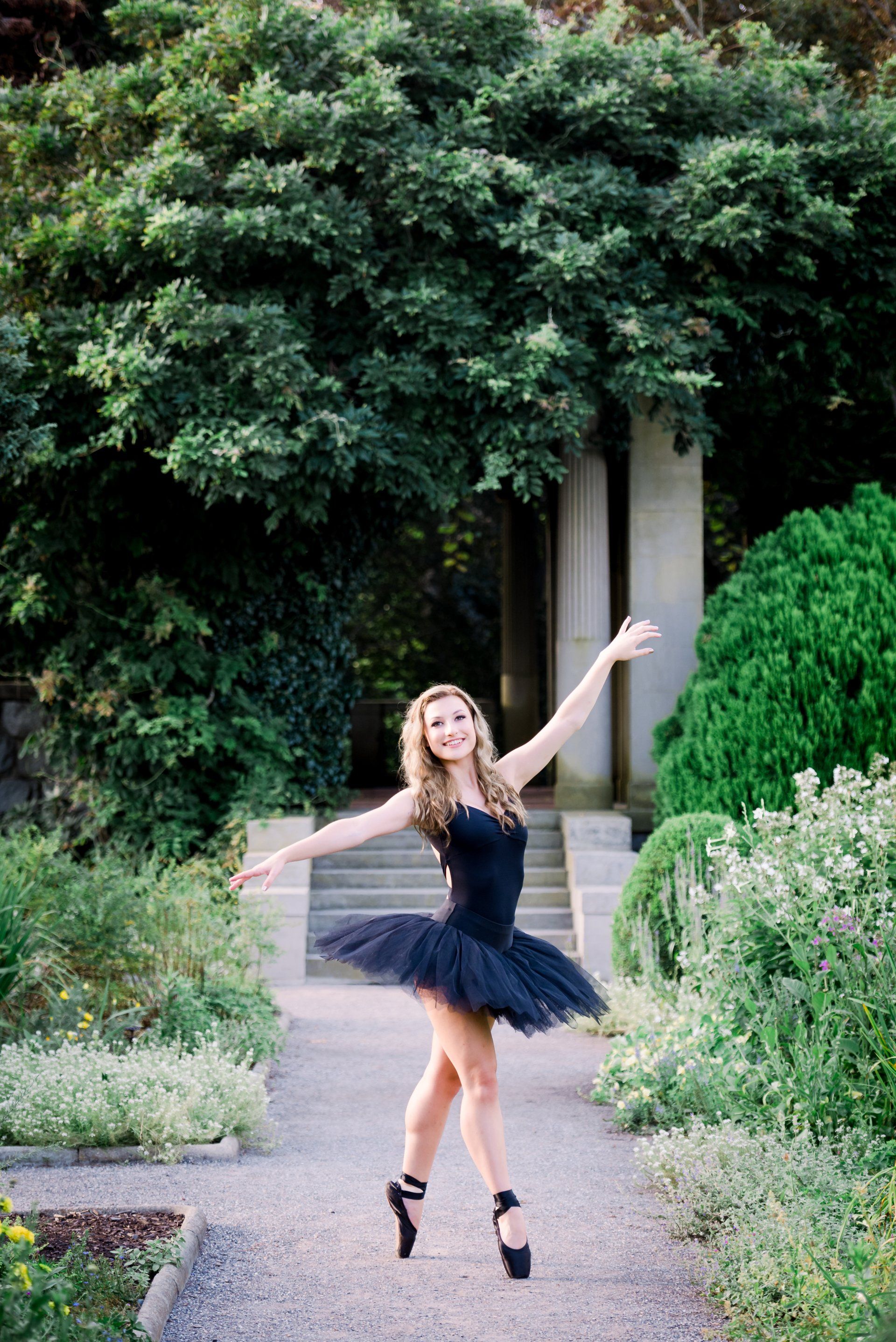 Woman in a black dress dancing on a garden path with trees and steps behind her