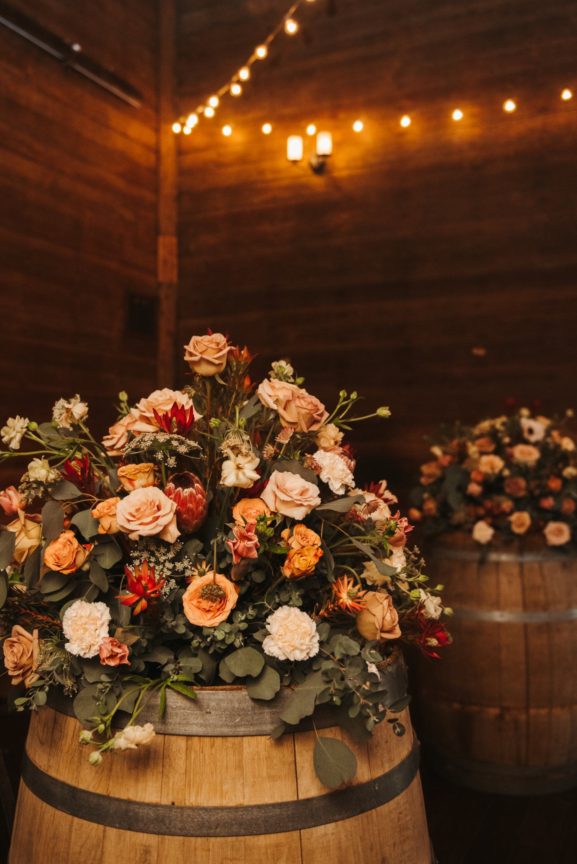 Flower arrangements in rustic wooden barrels under warm string lights in a barn-like venue