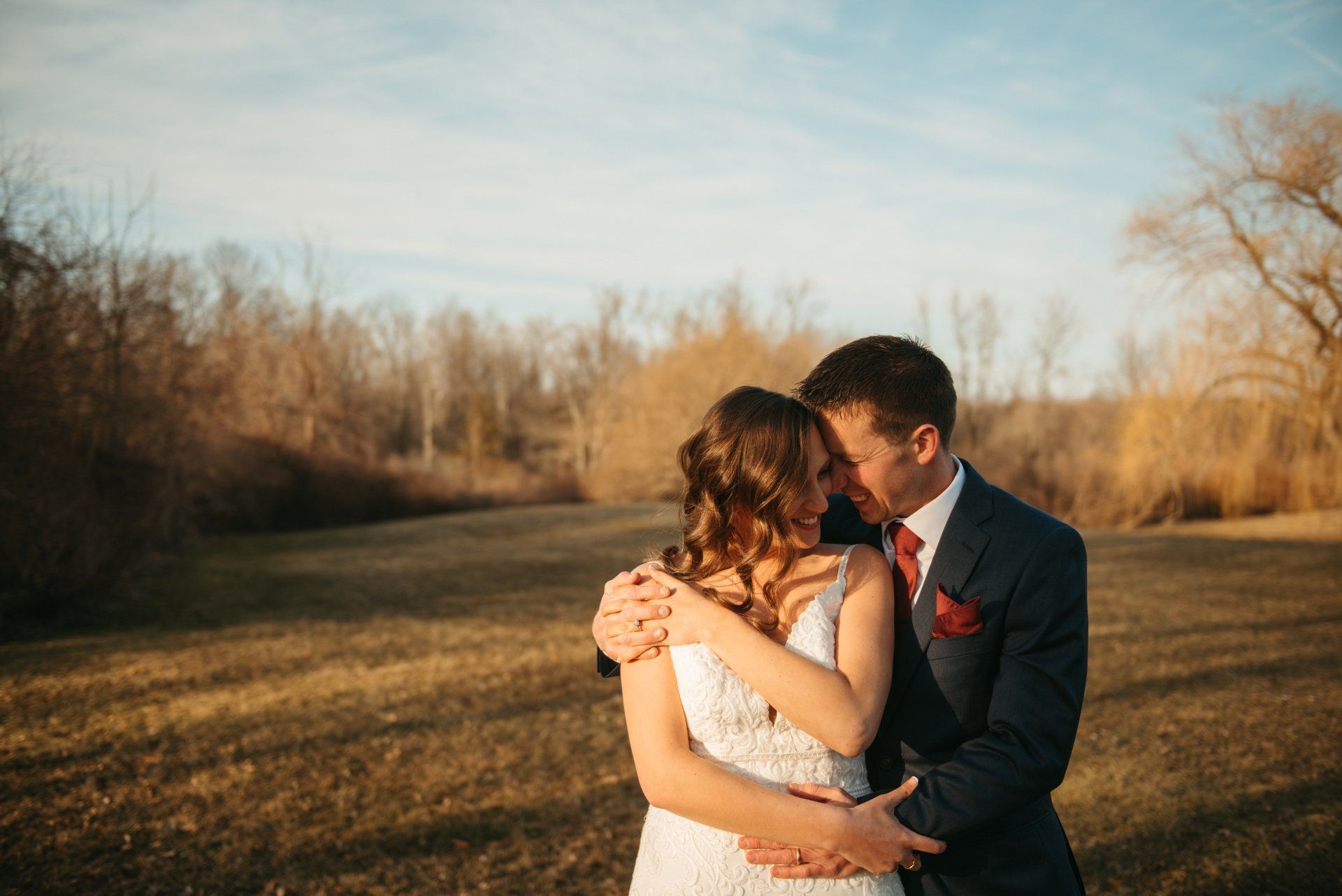 Couple embracing outdoors in a field at sunset, the woman in a white dress and the man in a dark suit.