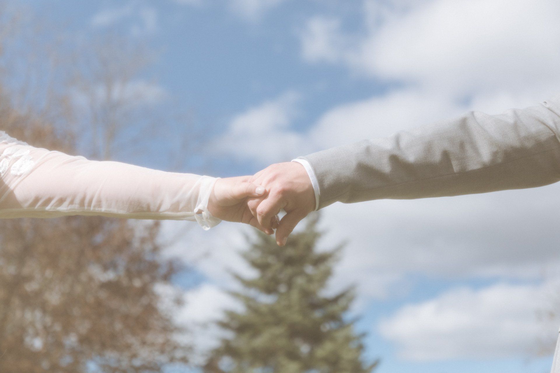 Two people holding hands outdoors against a blue sky with clouds and trees