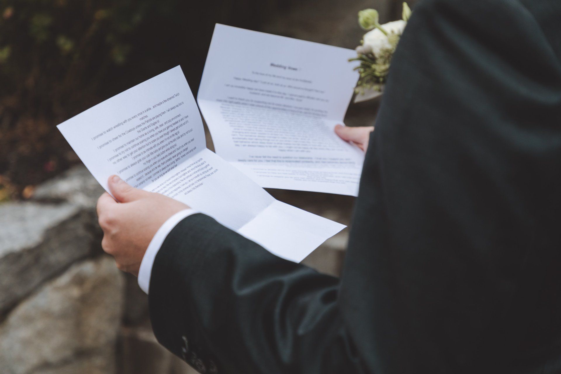 Person in a black suit holding several printed documents outdoors, with flowers in the background