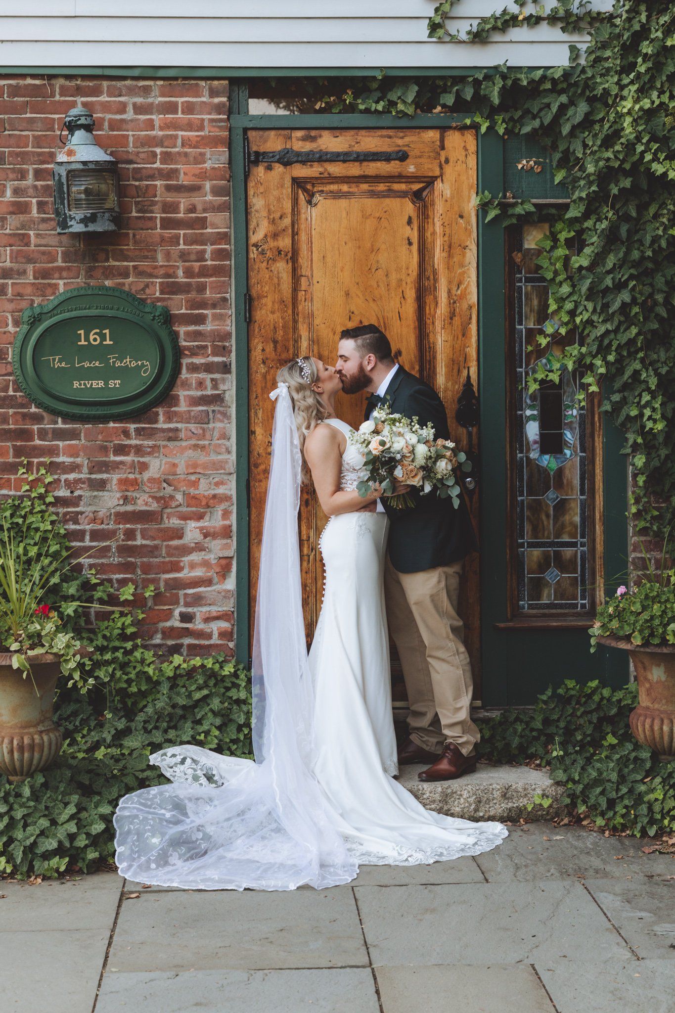 Bride and groom kissing outside a brick doorway, with a long white veil and bouquet.