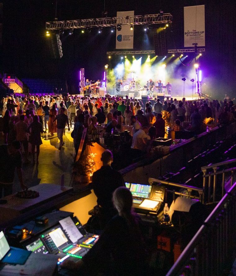 Crowded concert floor with bright stage lights and a soundboard in the foreground