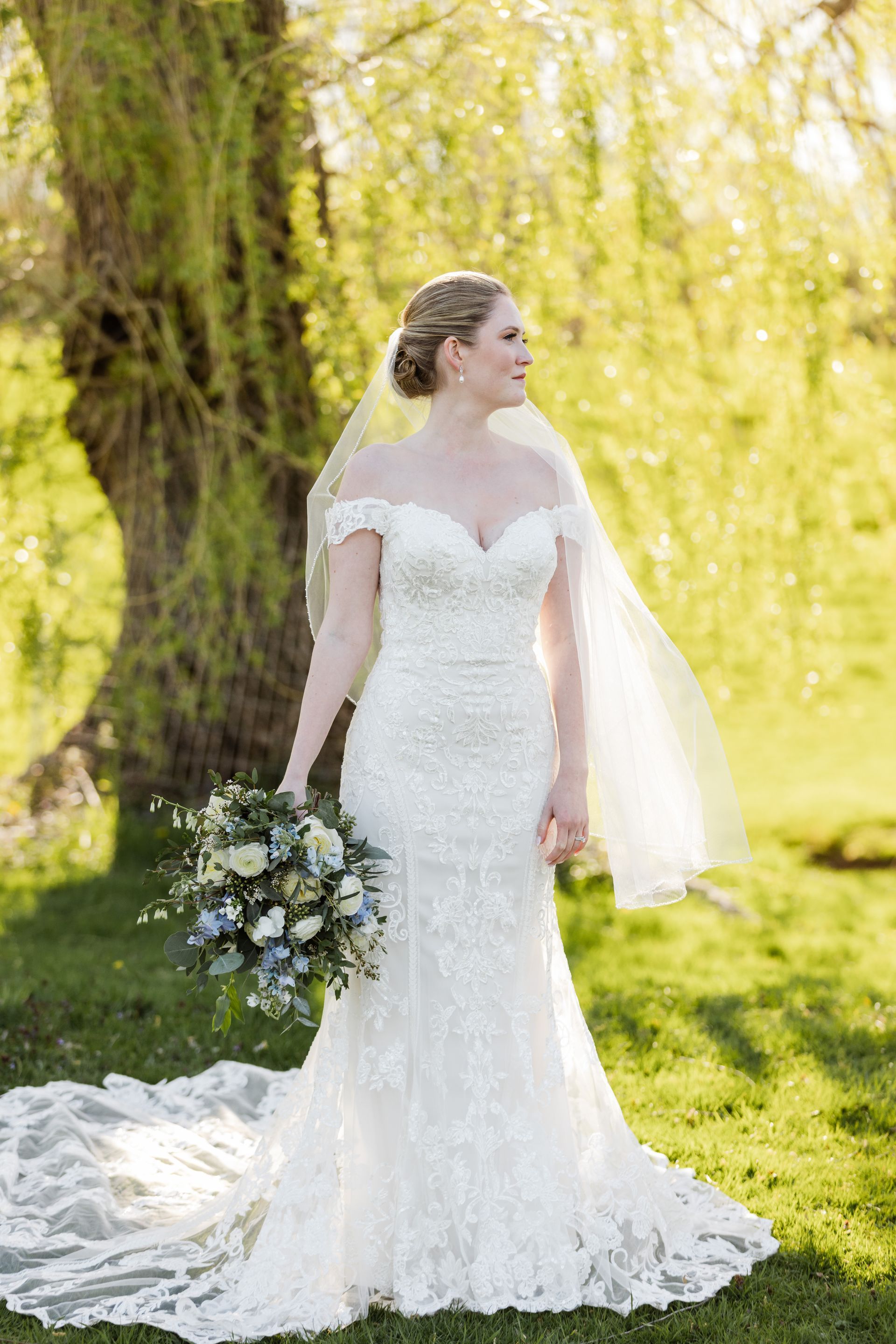 Bride in a white lace wedding dress holding a bouquet under a willow tree in a sunlit garden