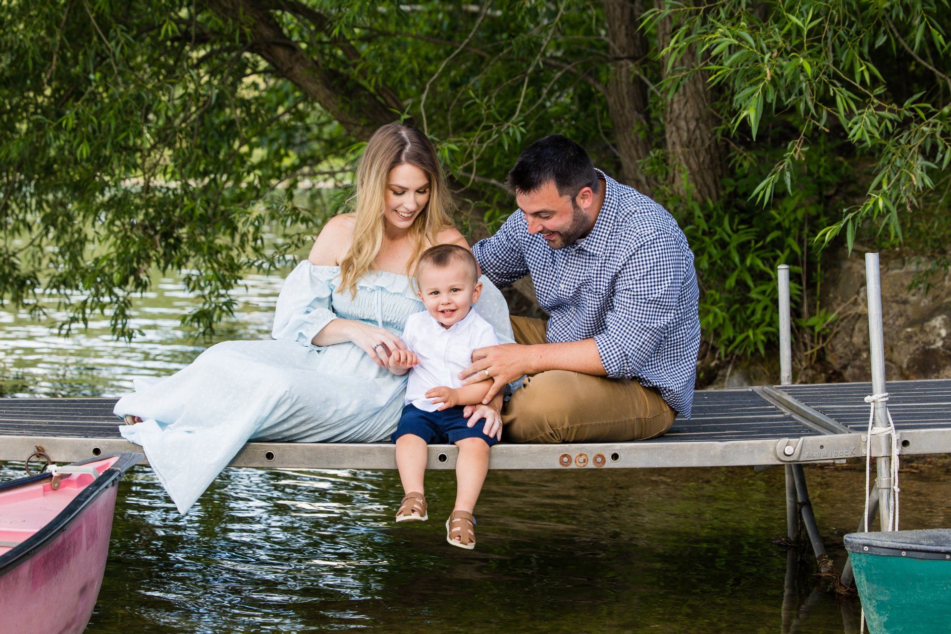 Family sitting on a dock by the water, smiling under green trees with a small child in the center