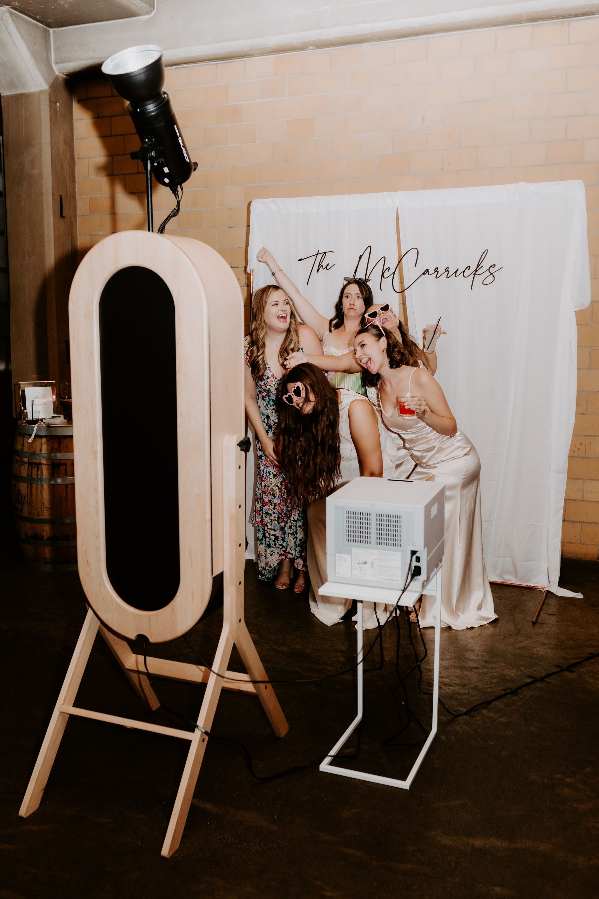 Four women posing in a photo booth setup with a white backdrop and oval mirror in a studio.