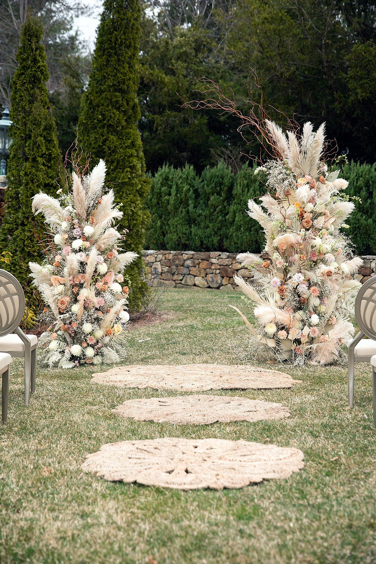Flower-lined garden aisle with tall cream and peach floral arrangements and round rug markers on the grass
