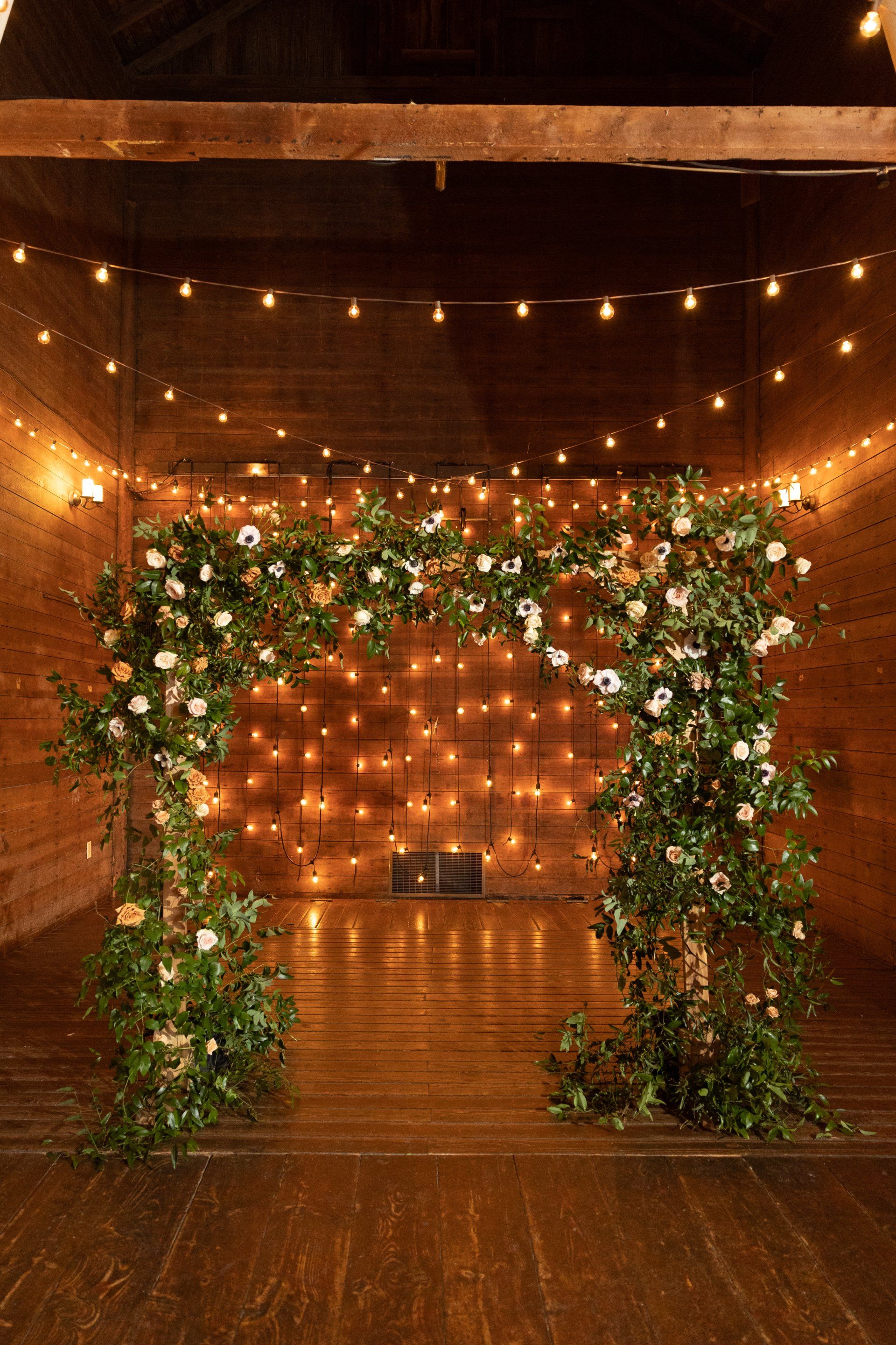 Floral arch at the end of a warmly lit wooden aisle with string lights in a rustic venue