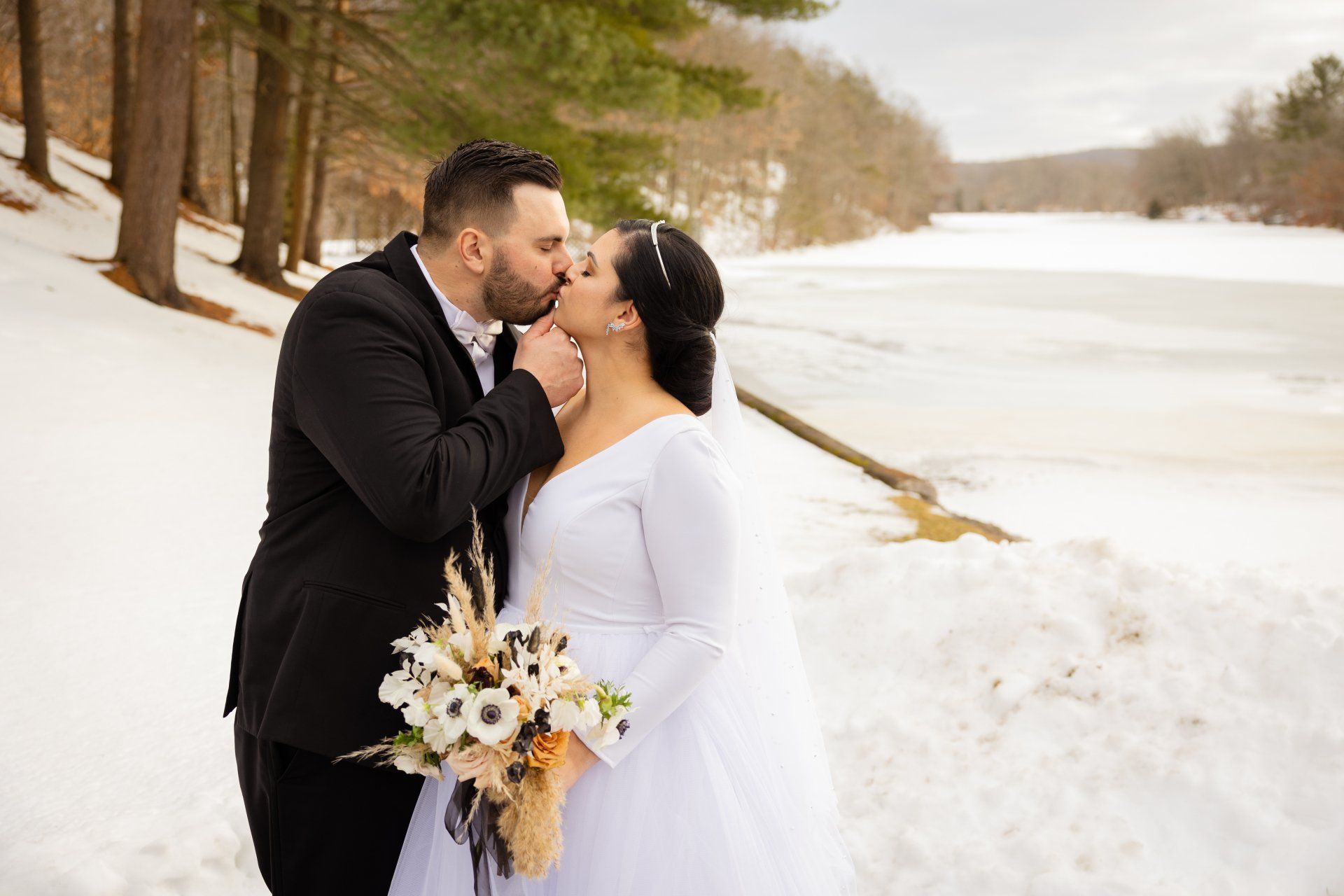 Couple kissing outdoors in winter by a snowy lakeshore, with a bouquet in hand